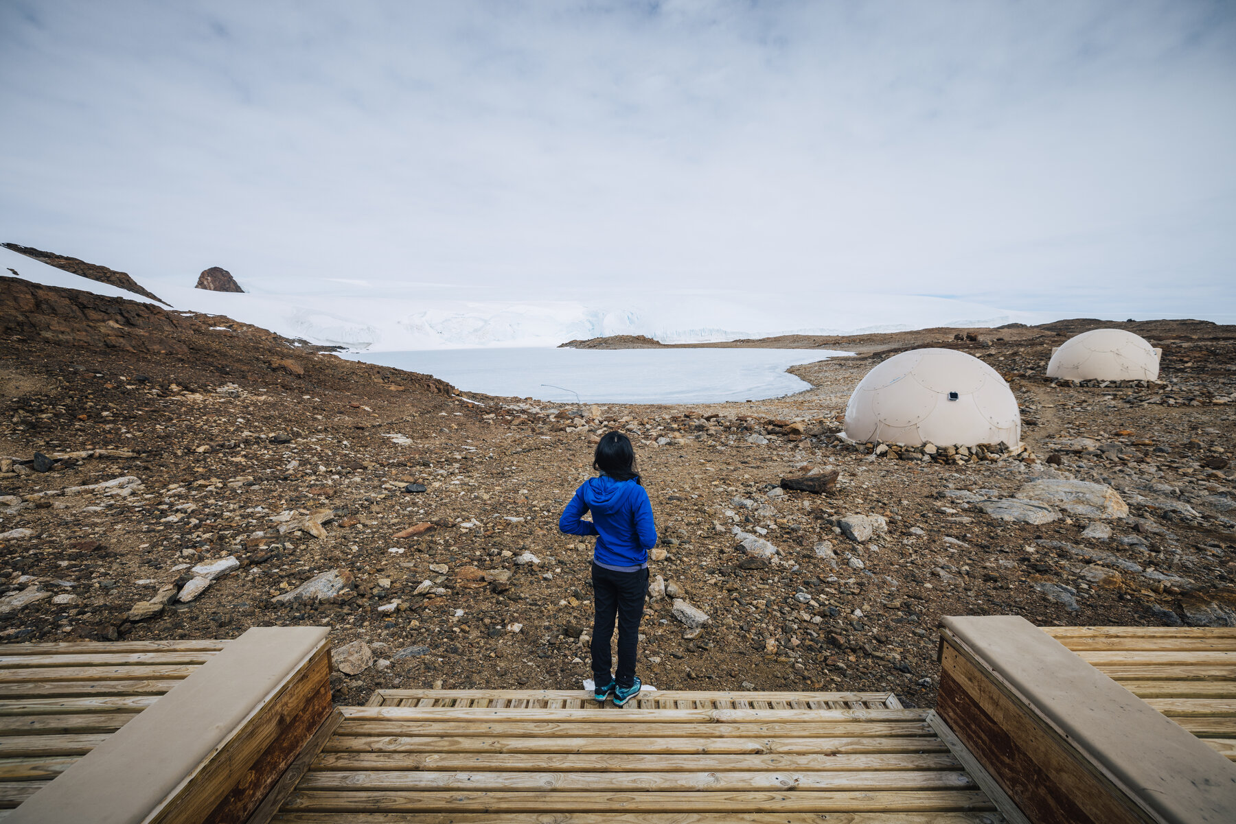 Grace in a blue parka standing at the edge of the wooden deck looking out across the frozen lake toward the pods and glacier wall