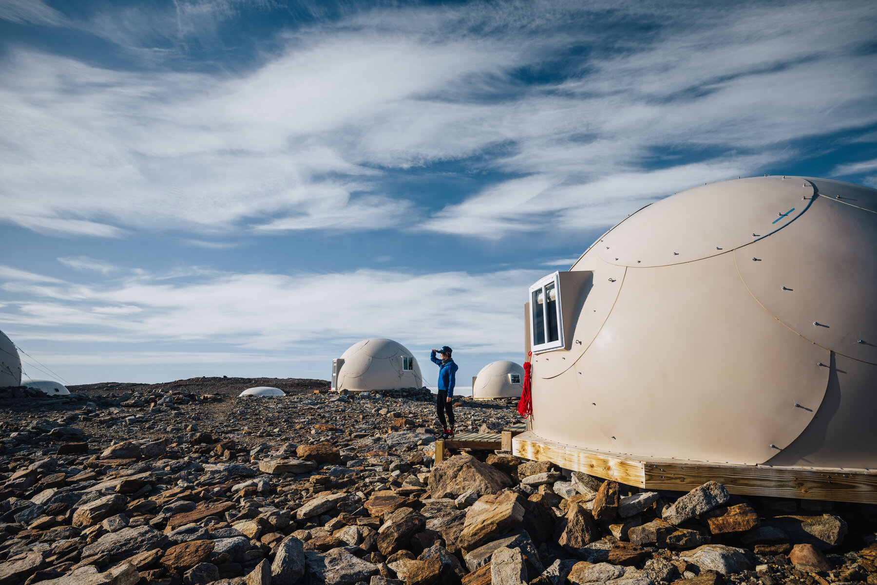 A white dome pod at Whichaway Camp with its circular window reflecting the sky, a figure in a blue parka on the wooden deck, and other pods and the frozen lake stretching into the distance