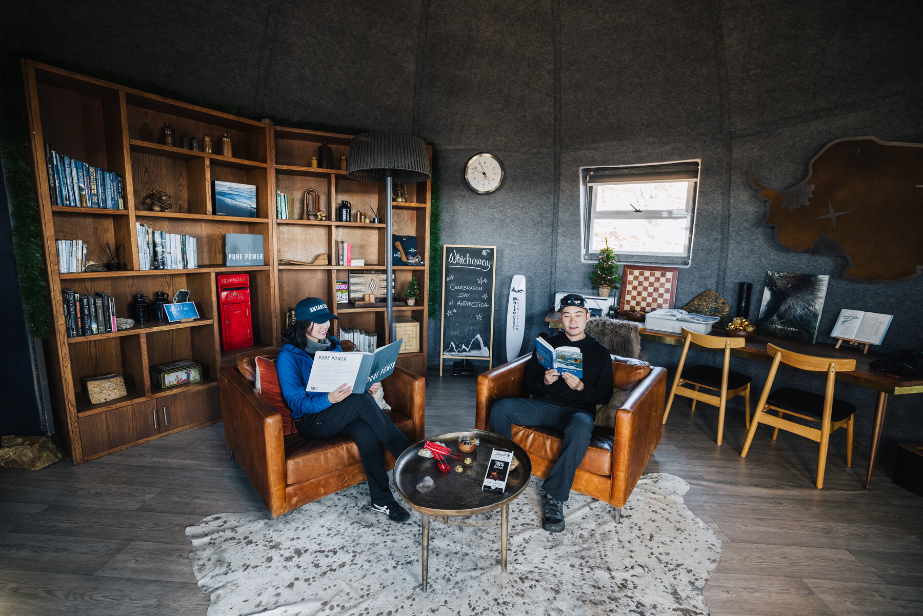 Jason and Grace reading in leather club chairs in the Whichaway library pod with wooden bookshelves, a chess table, and polar-themed art
