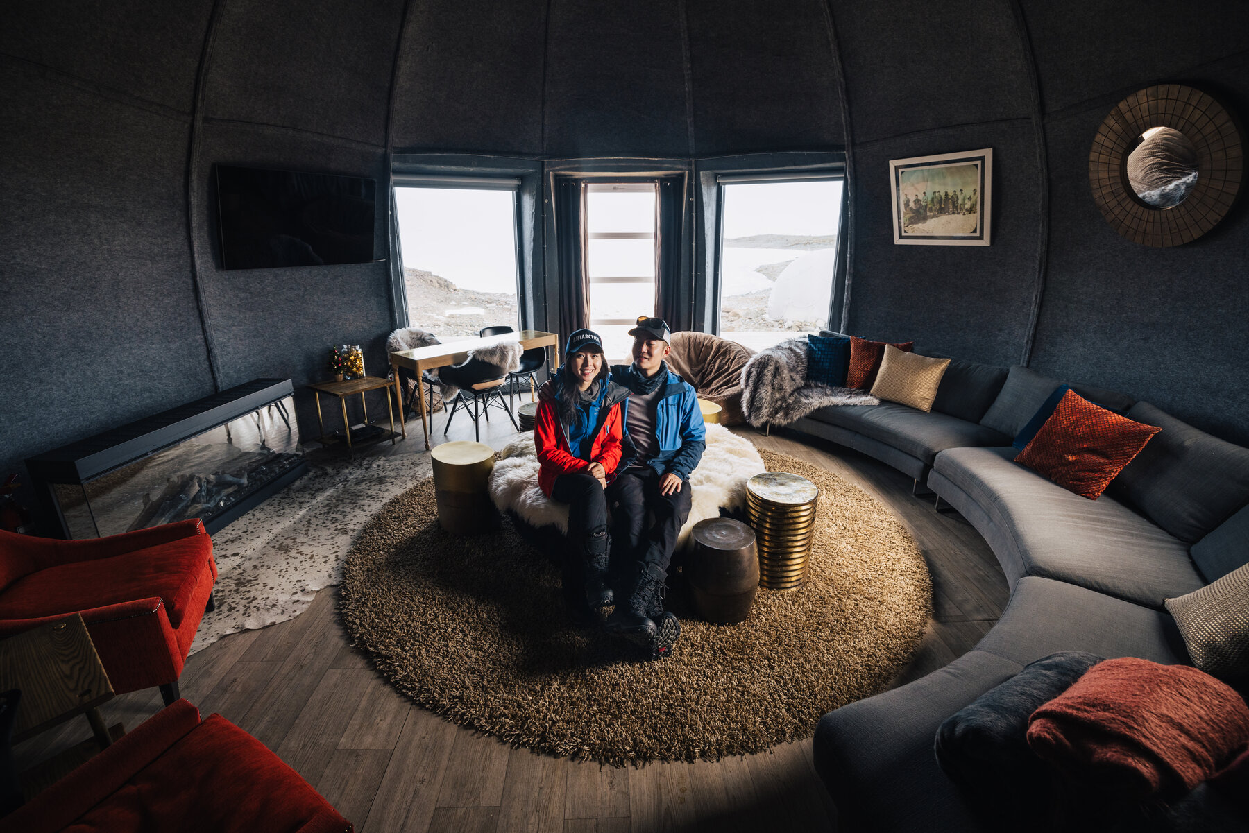 Jason and Grace seated together on a round sheepskin-covered ottoman in the main lounge pod, with a curved grey sofa, rugs, and large windows overlooking the polar landscape