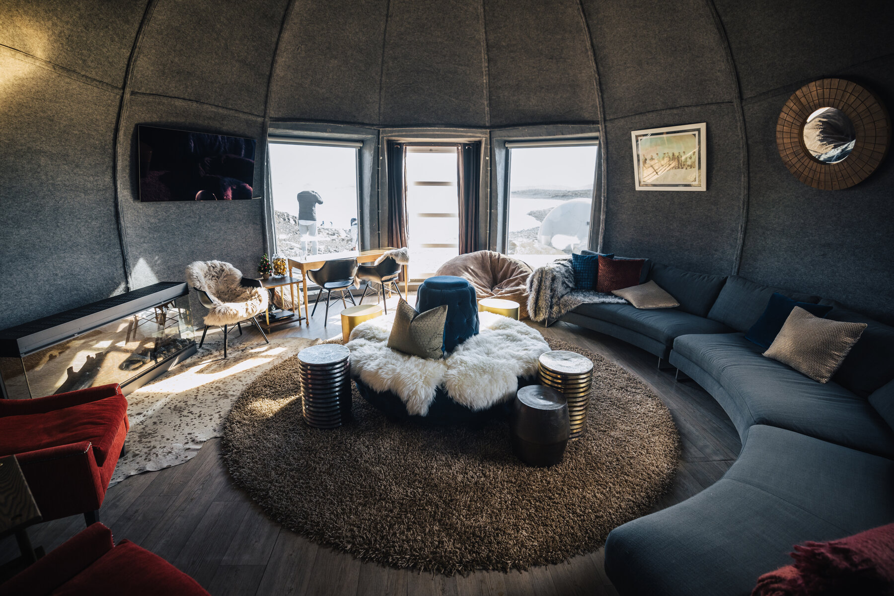 Wide view of the Whichaway main lounge pod with curved grey sectional sofa, round sheepskin ottoman, rugs, and tall windows looking onto the ice
