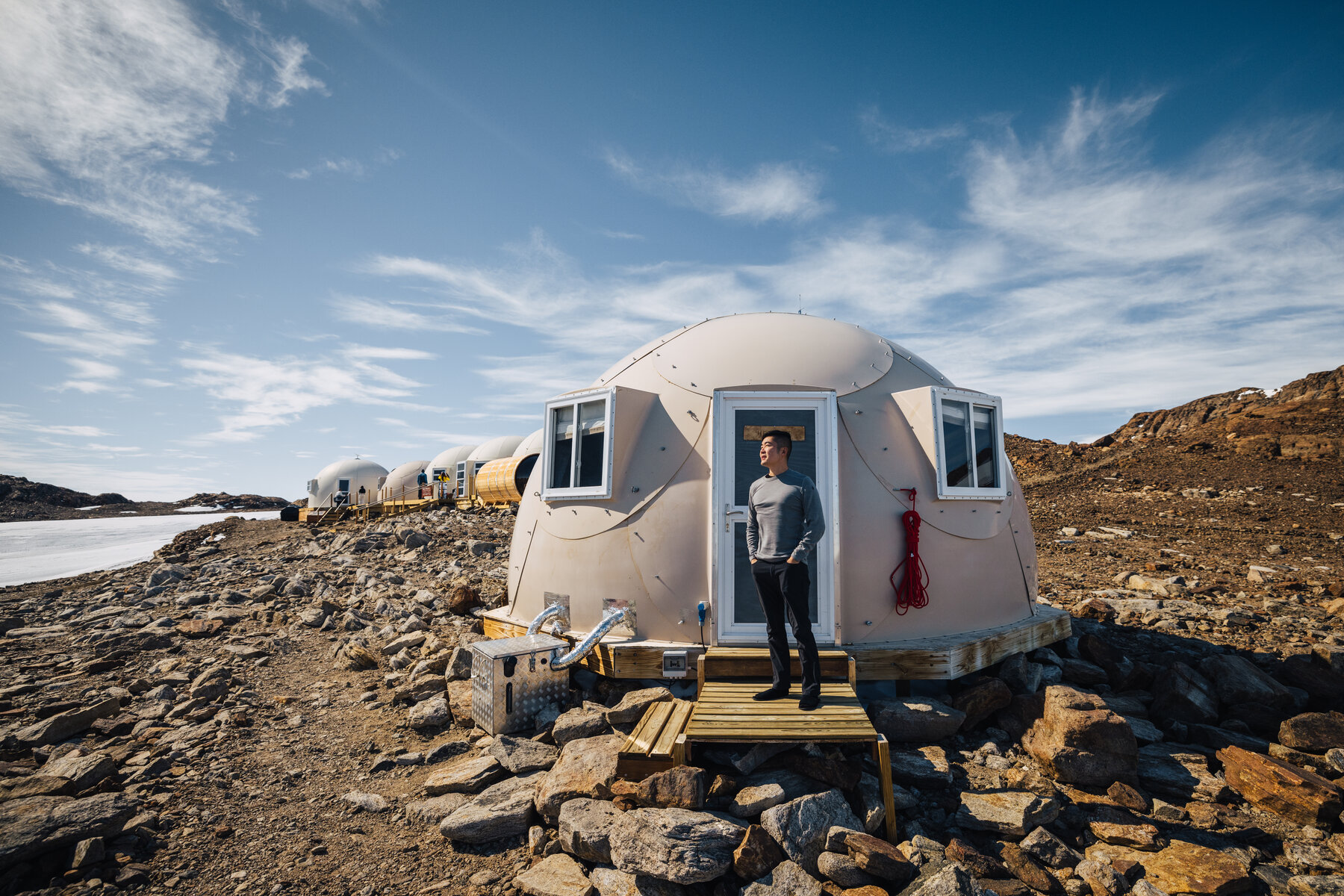 Jason in a grey shirt standing on the wooden deck in the open doorway of a Whichaway pod with the row of other pods and the frozen lake behind