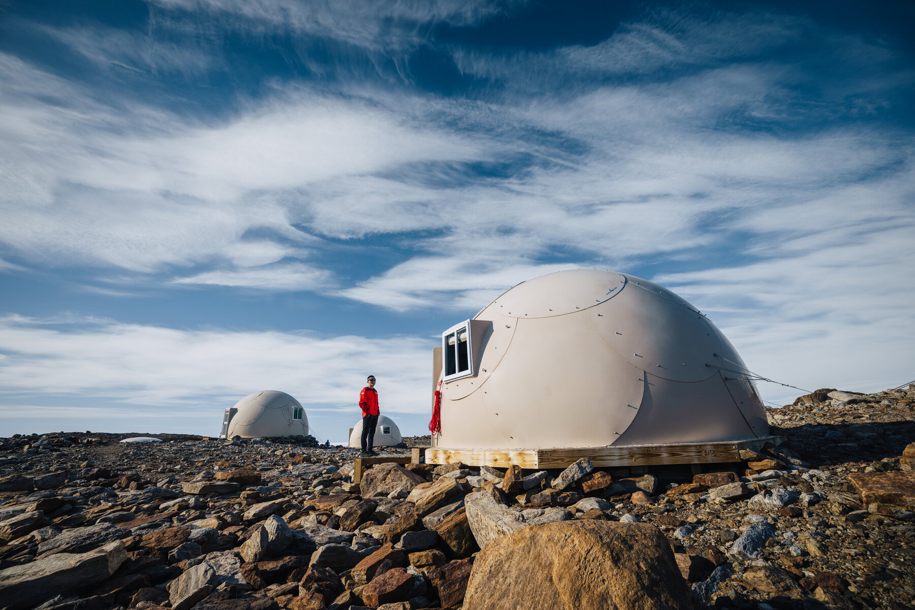 Figure in a red parka standing between two white dome pods on the Whichaway rocky outcrop with swept cirrus clouds overhead