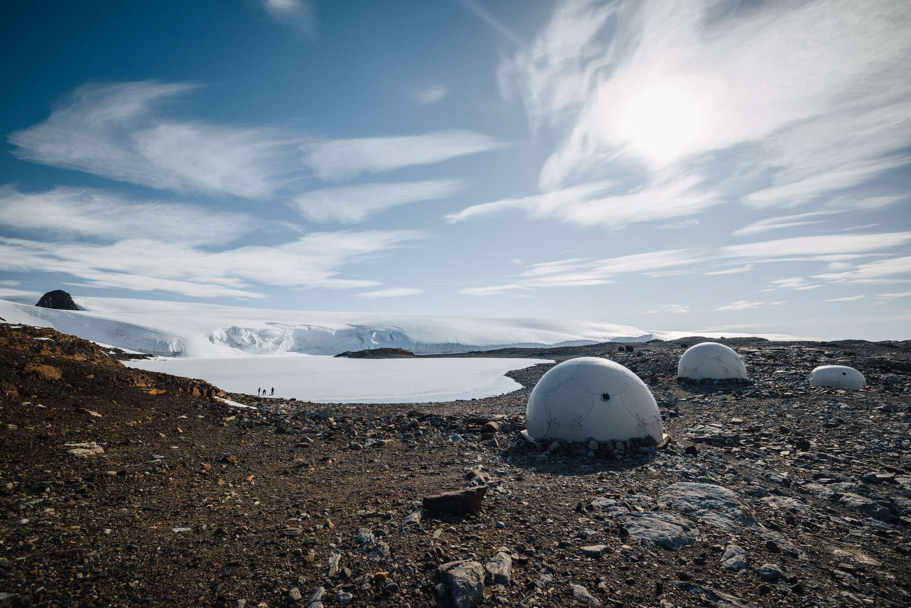 Wide panorama of the Whichaway Camp pods spread across the dark rocky outcrop with the frozen lake and glacier wall stretching to the horizon behind