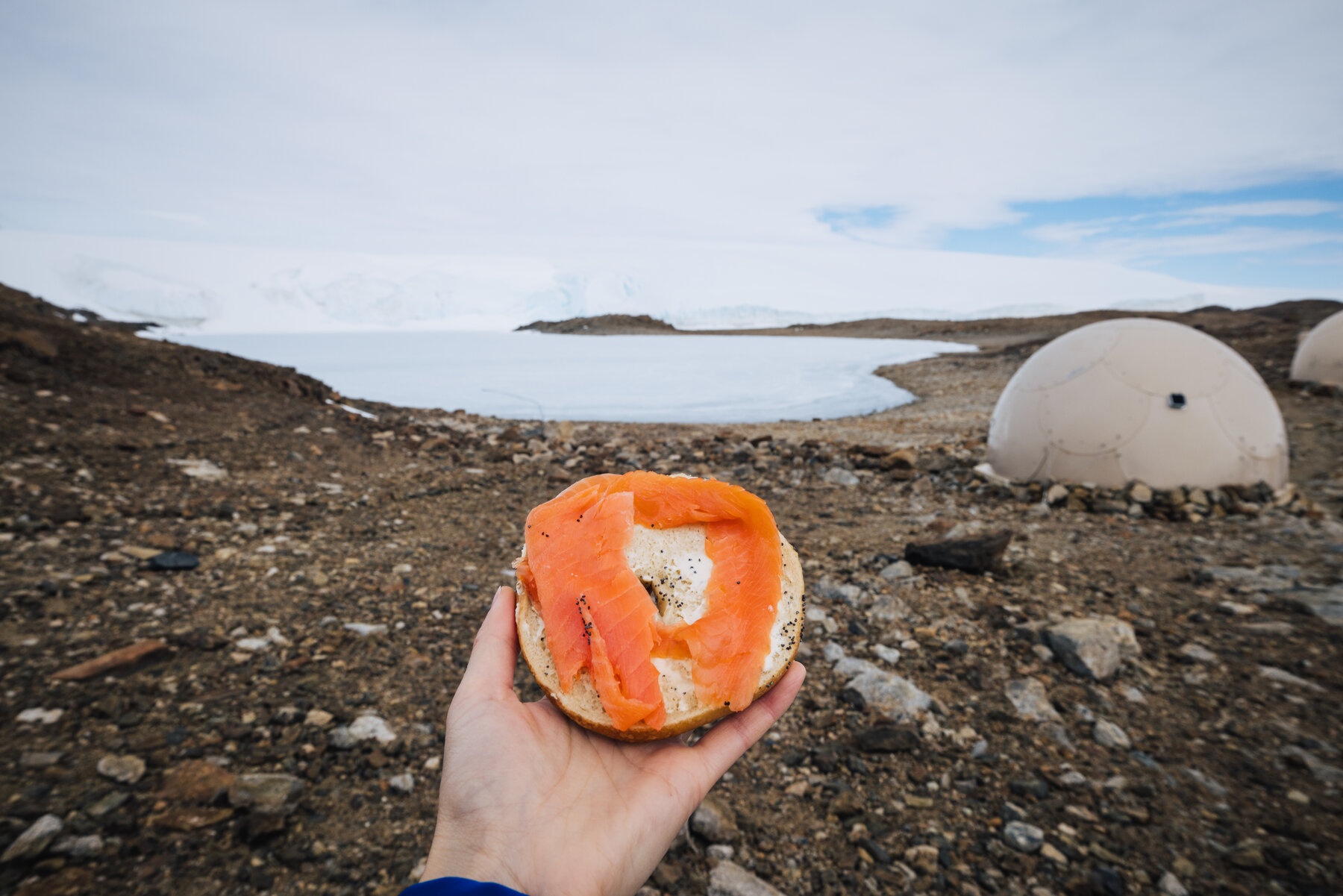 Hand holding a smoked salmon bagel with the Whichaway dome pods and frozen lake in the background