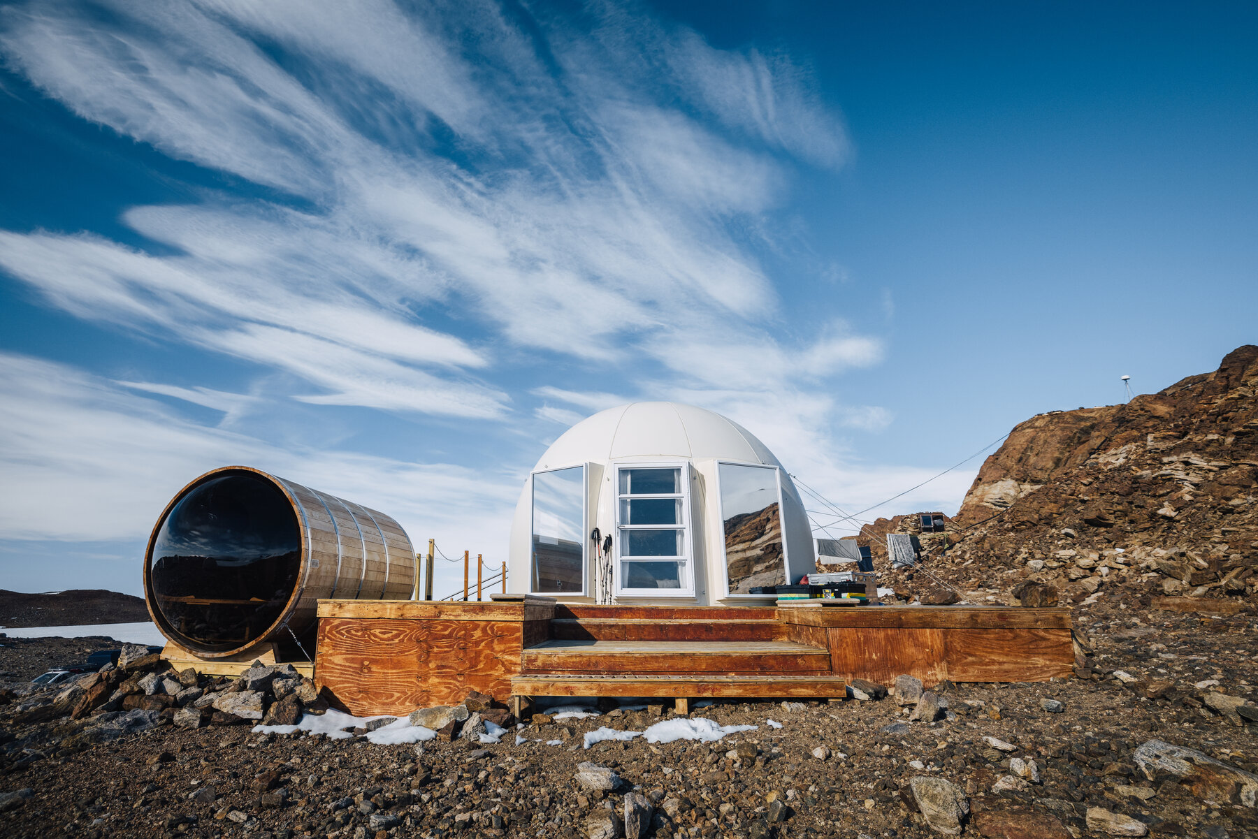 Exterior of the cylindrical wooden cedar sauna next to a white dome pod at Whichaway, set on a rocky outcrop with sweeping cirrus clouds overhead