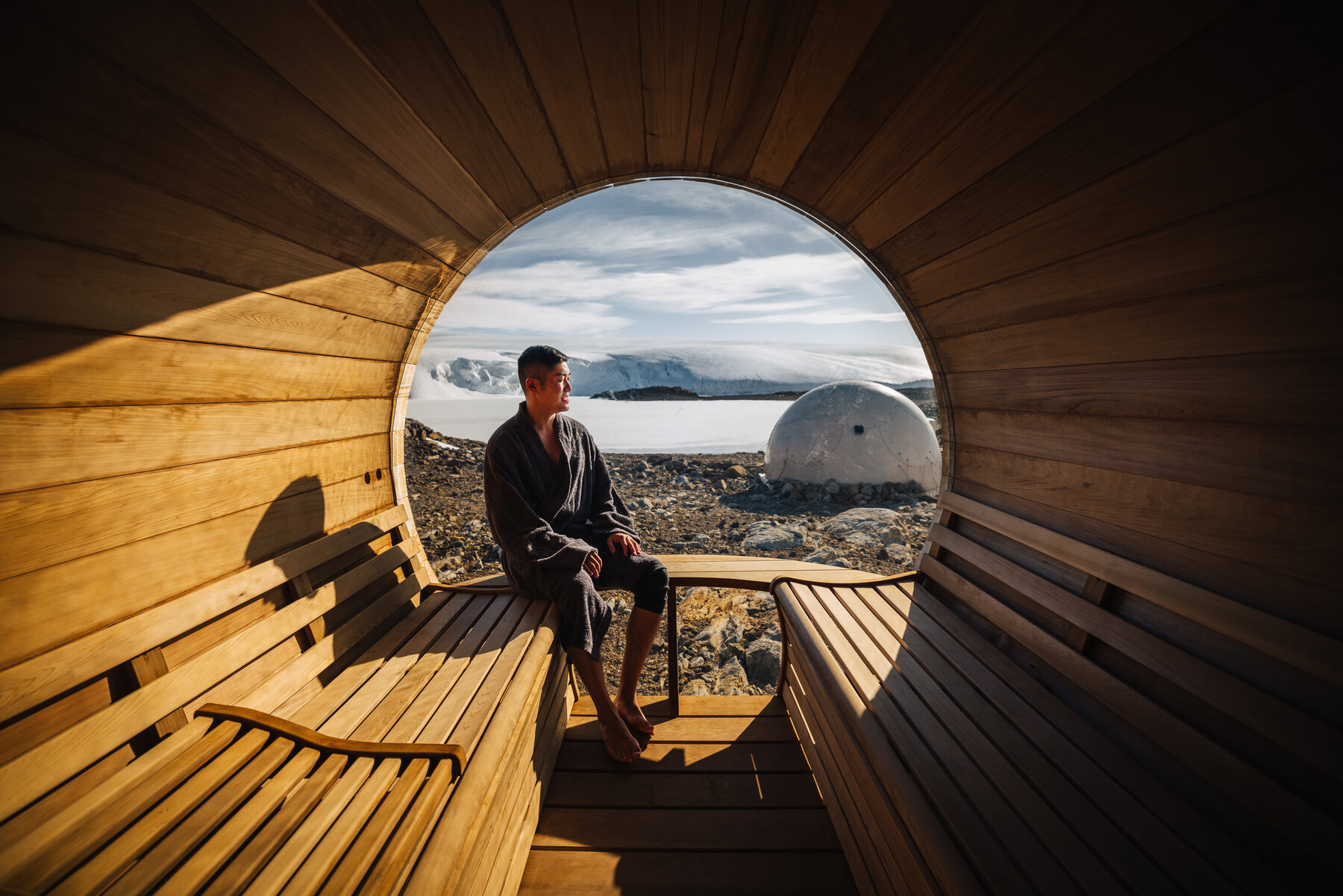 Jason in a dark robe seated inside the wooden cedar sauna looking out through its large circular window at a dome pod and the glacier beyond