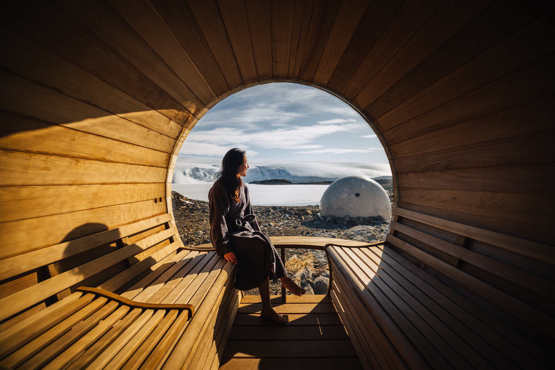 Grace in a dark robe seated inside the wooden cedar sauna looking out through its large circular window at a dome pod and the glacier beyond
