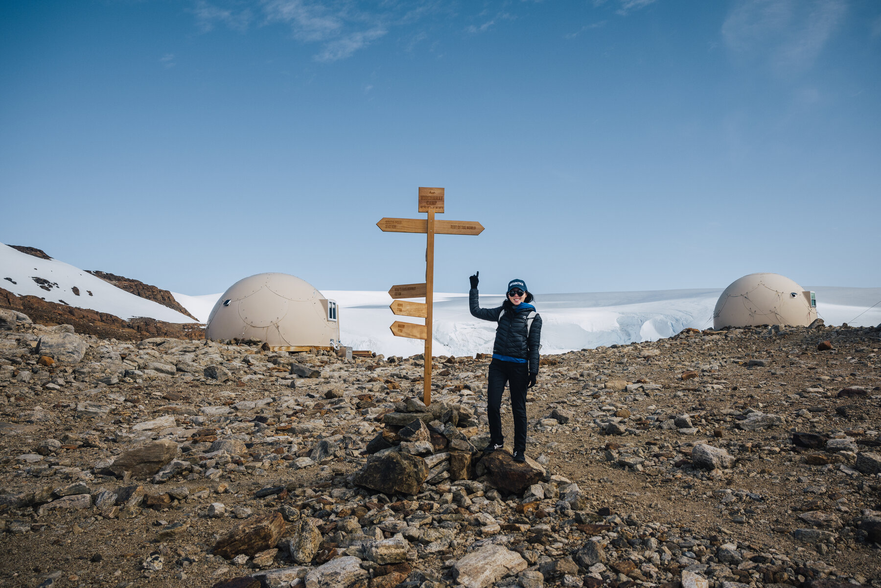 Grace waving beside the wooden Whichaway directional signpost between two pods with the glacier behind