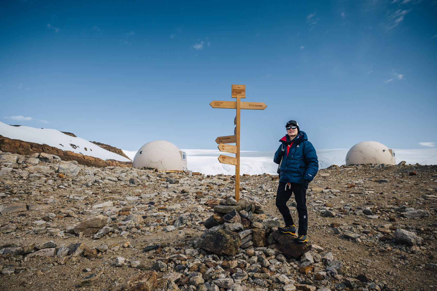 Jason standing beside the wooden Whichaway directional signpost between two pods with the glacier in the distance