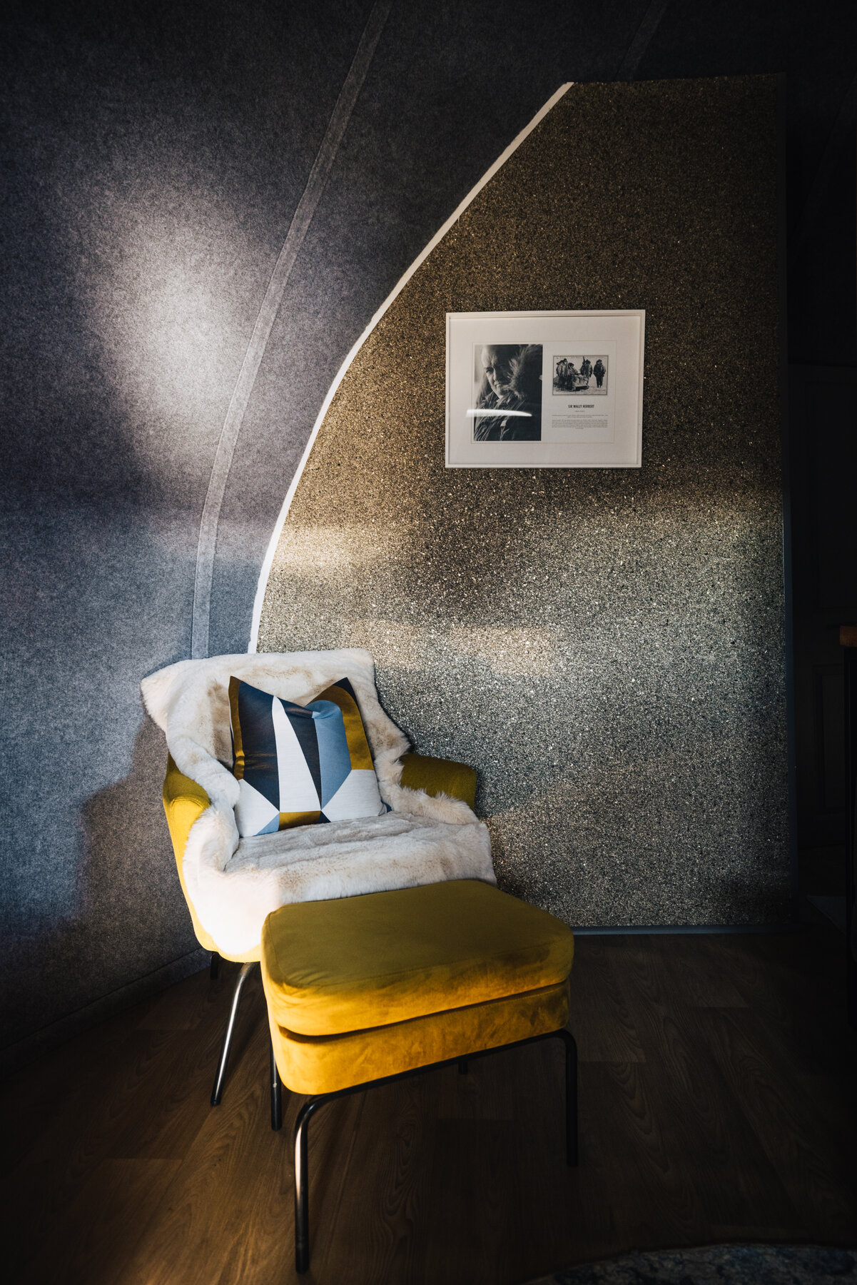Yellow velvet armchair and ottoman in a Whichaway pod reading nook with a framed black-and-white photograph on the felt-textured wall