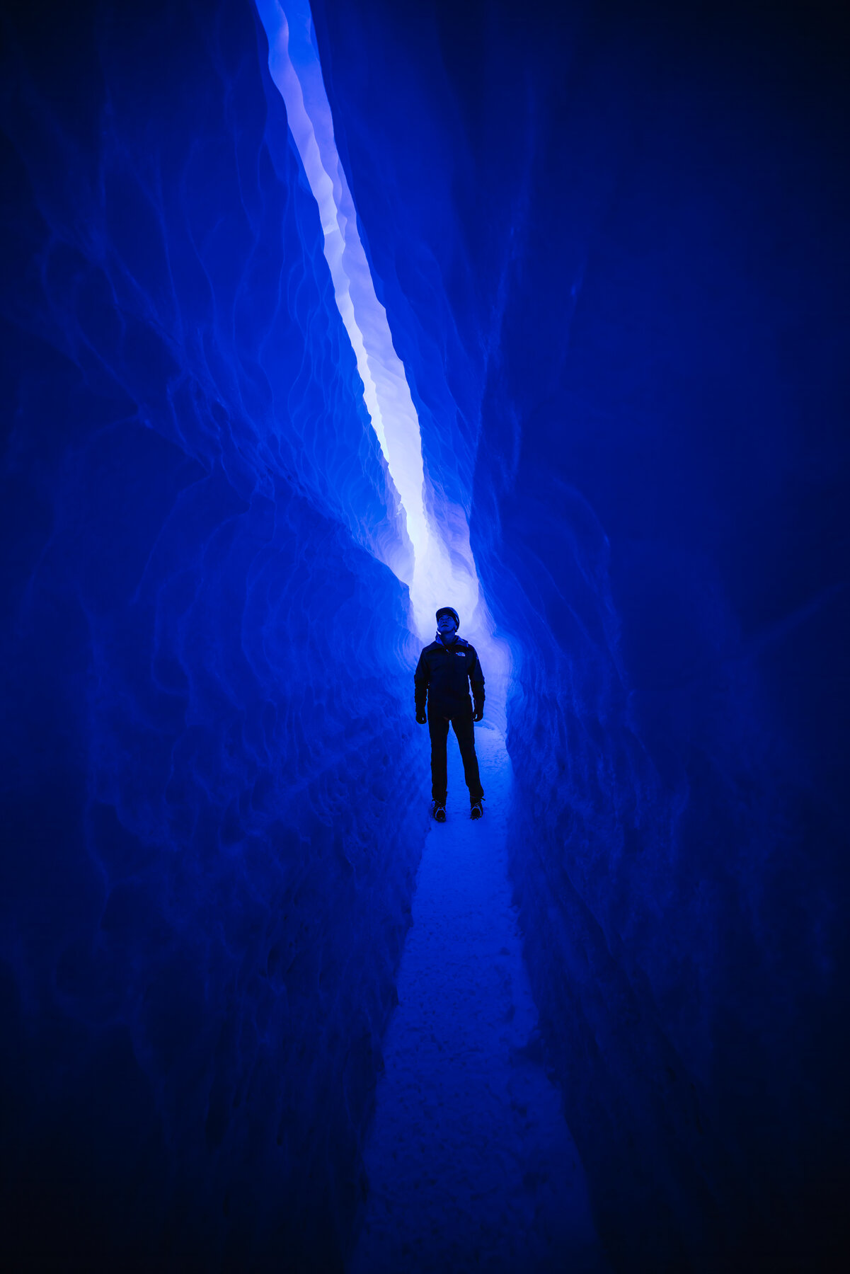 Silhouette of a climber framed in the narrow opening of a deep blue glacial crevasse with luminous ice walls curving inward