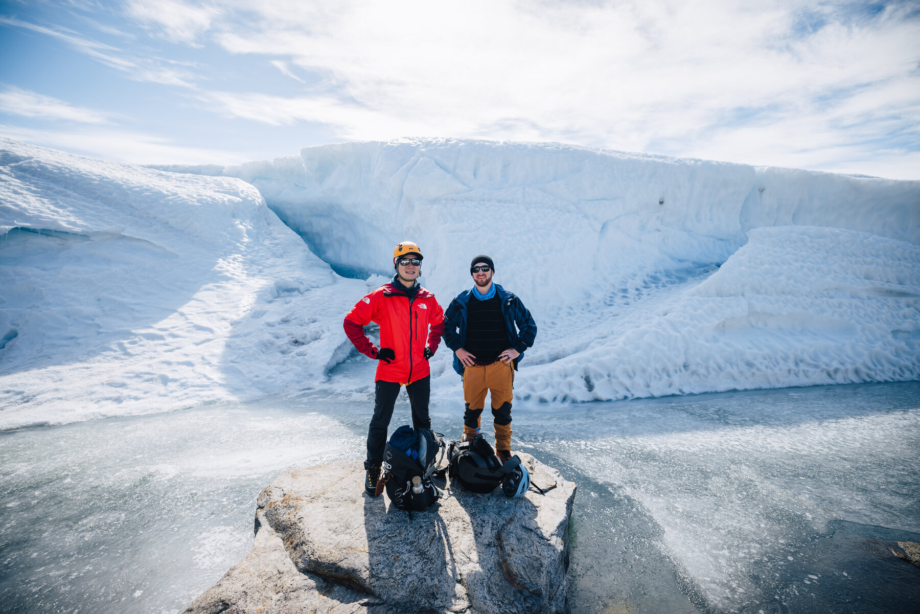 Jason in a red jacket and a companion standing with their packs at the foot of a pale blue ice wall under a bright sky