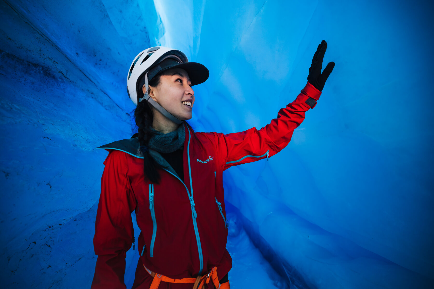 Grace in a red jacket and white climbing helmet smiling inside the luminous blue ice tunnel