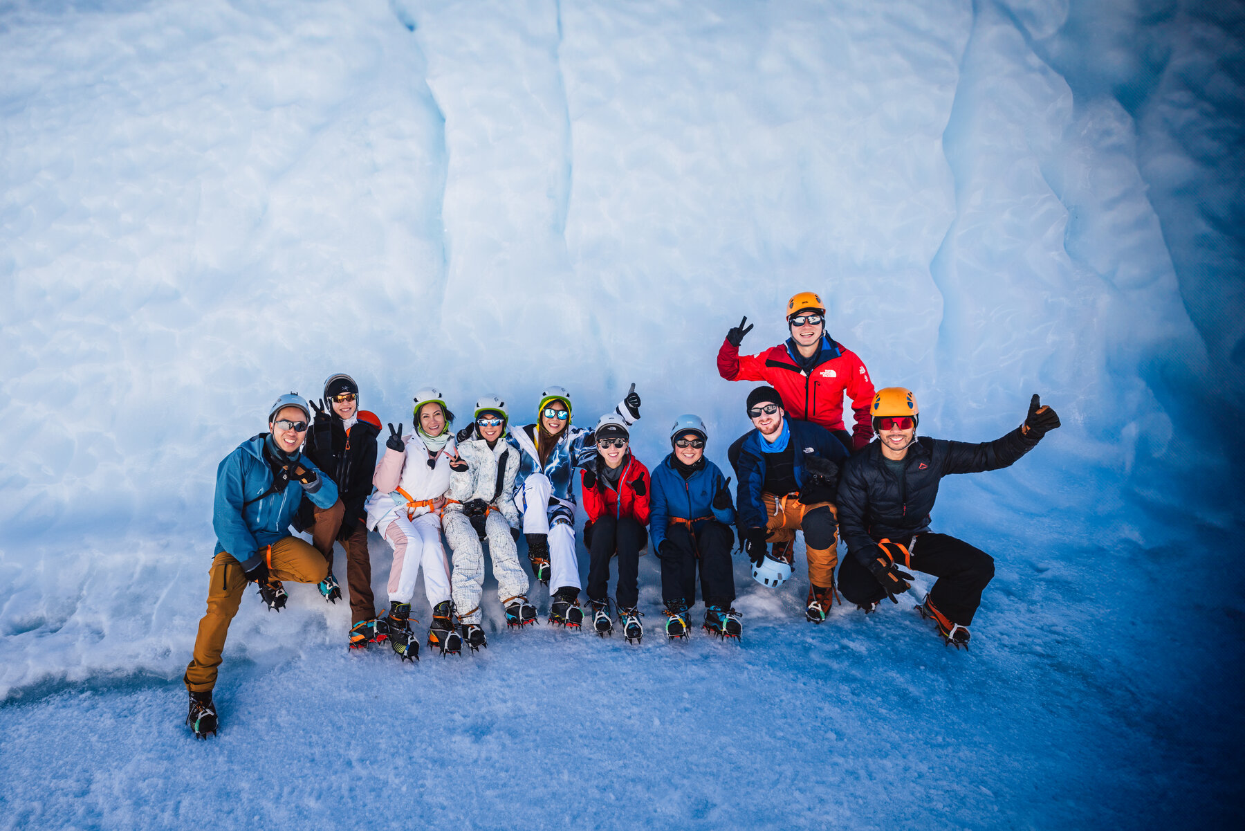 Group of travelers in climbing helmets and crampons posing together at the foot of a pale blue ice wall