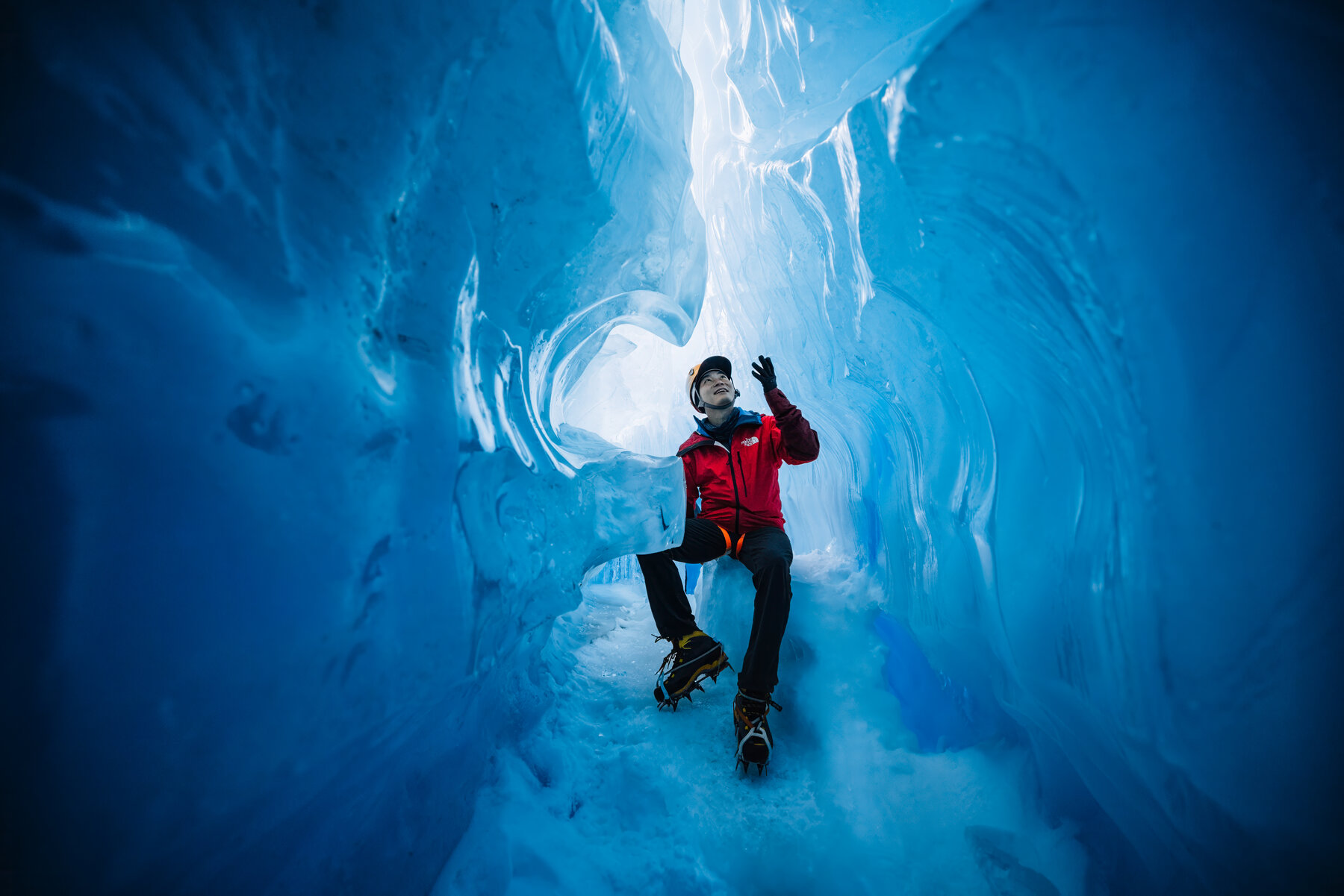 Jason in a red parka and yellow climbing helmet reaching up to touch the luminous blue ceiling of a glacial ice tunnel