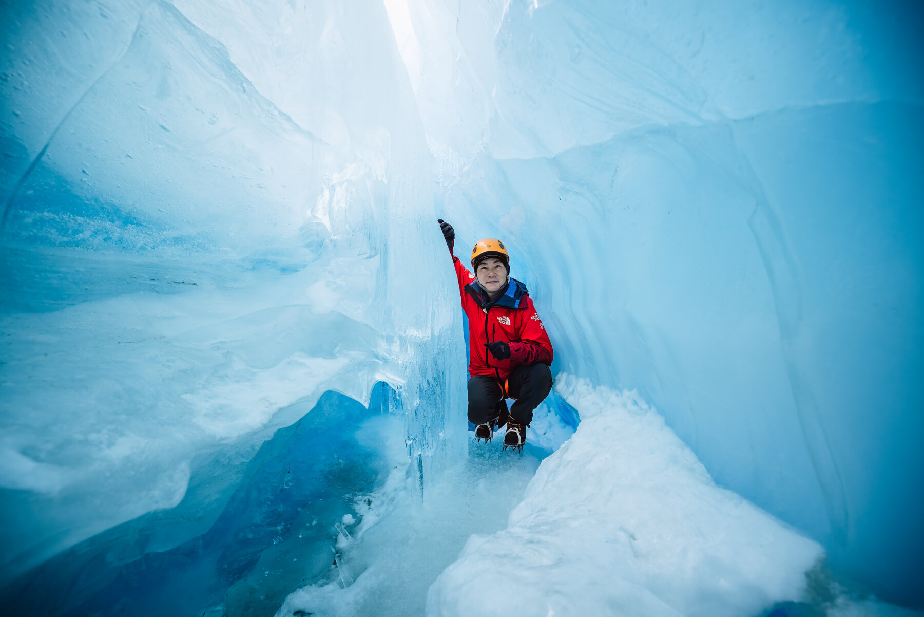 Jason in a yellow climbing helmet and red parka reaching up to touch the blue ice ceiling of a glacial tunnel