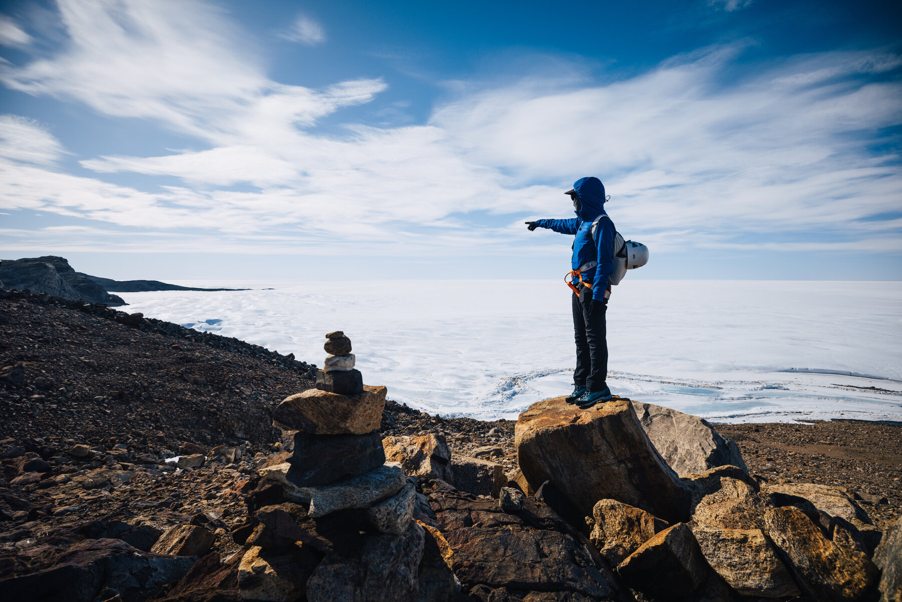 Grace standing on a boulder beside a small stone cairn pointing out across the vast ice sheet under a deep blue Antarctic sky