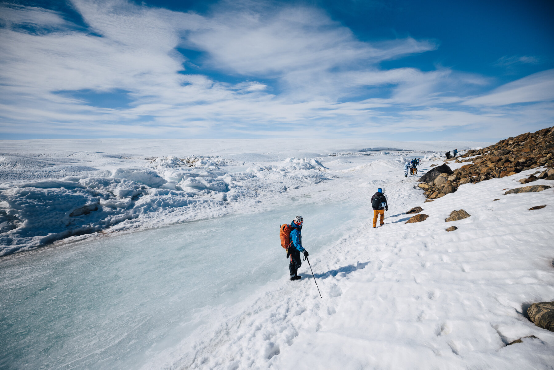 Group of parka-clad travelers hiking in single file across a flat ice plain toward distant ice wave formations