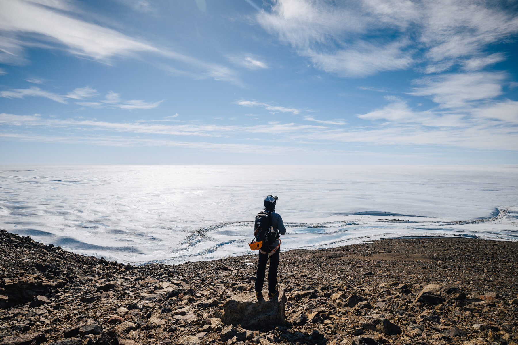 Lone figure in dark clothing standing on a rocky ridge overlooking the vast ice sheet stretching toward the horizon