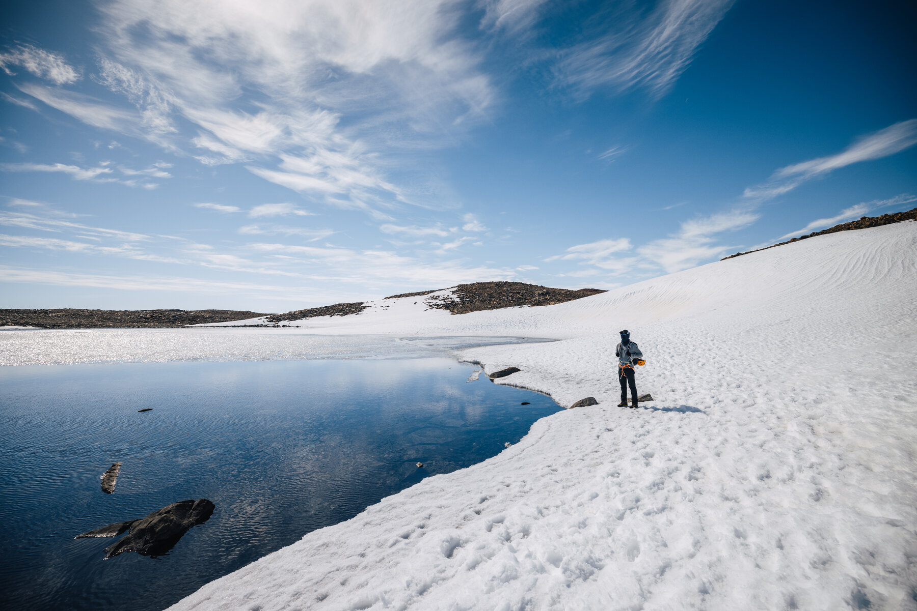 Deep-blue meltwater pond set into the snow-covered plateau with a lone figure on the bank and rocky outcrops behind