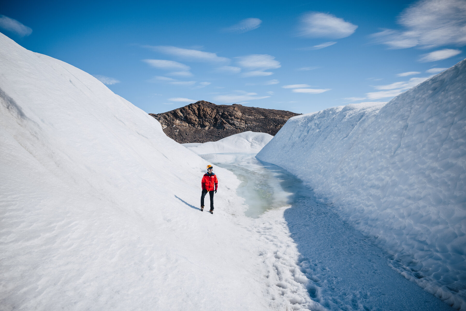 Figure in a red jacket walking through a snow canyon with a frozen meltwater channel running along its floor and a rocky ridge beyond