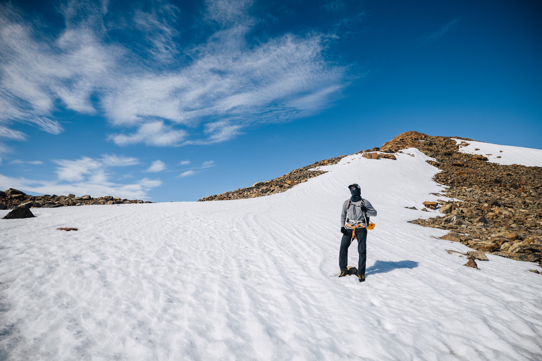 Solo climber in a light grey jacket and harness standing on a snow slope below a rocky ridge