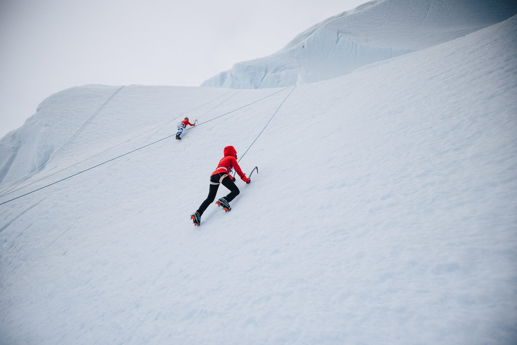 Two climbers in red jackets ascending a steep Antarctic snow slope with crampons and ice axes, ropes running between them