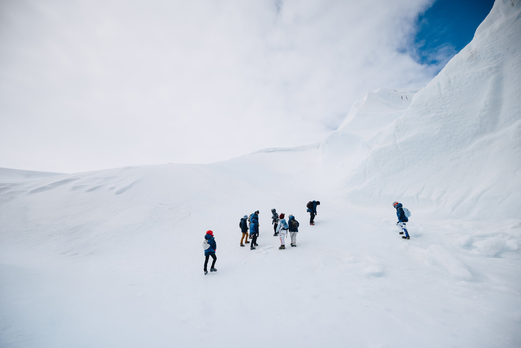 Group of climbers in winter gear hiking up a snowfield toward the steep flanks of the nunatak