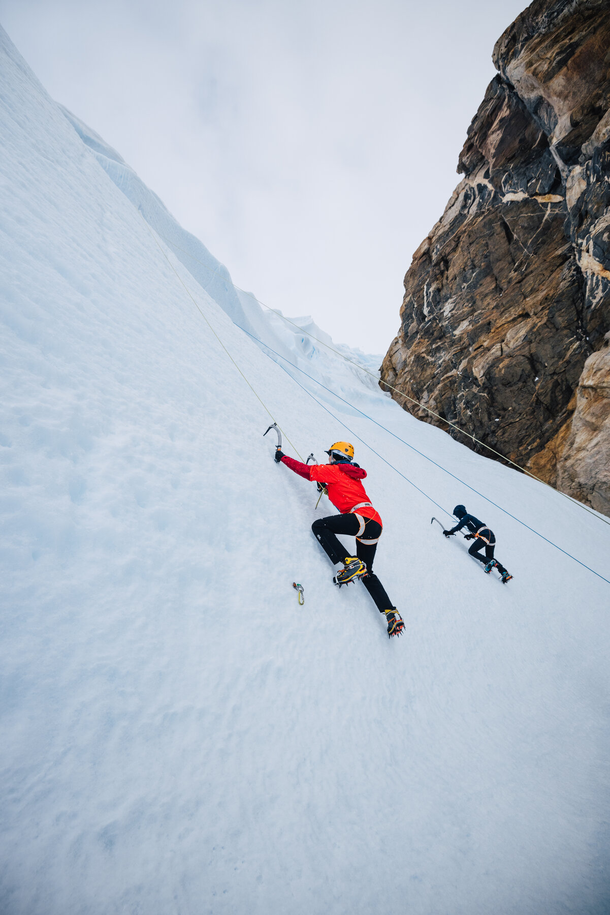 Climber in red jacket and yellow helmet ascending a steep blue ice wall using ice axes and crampons with a belayer below