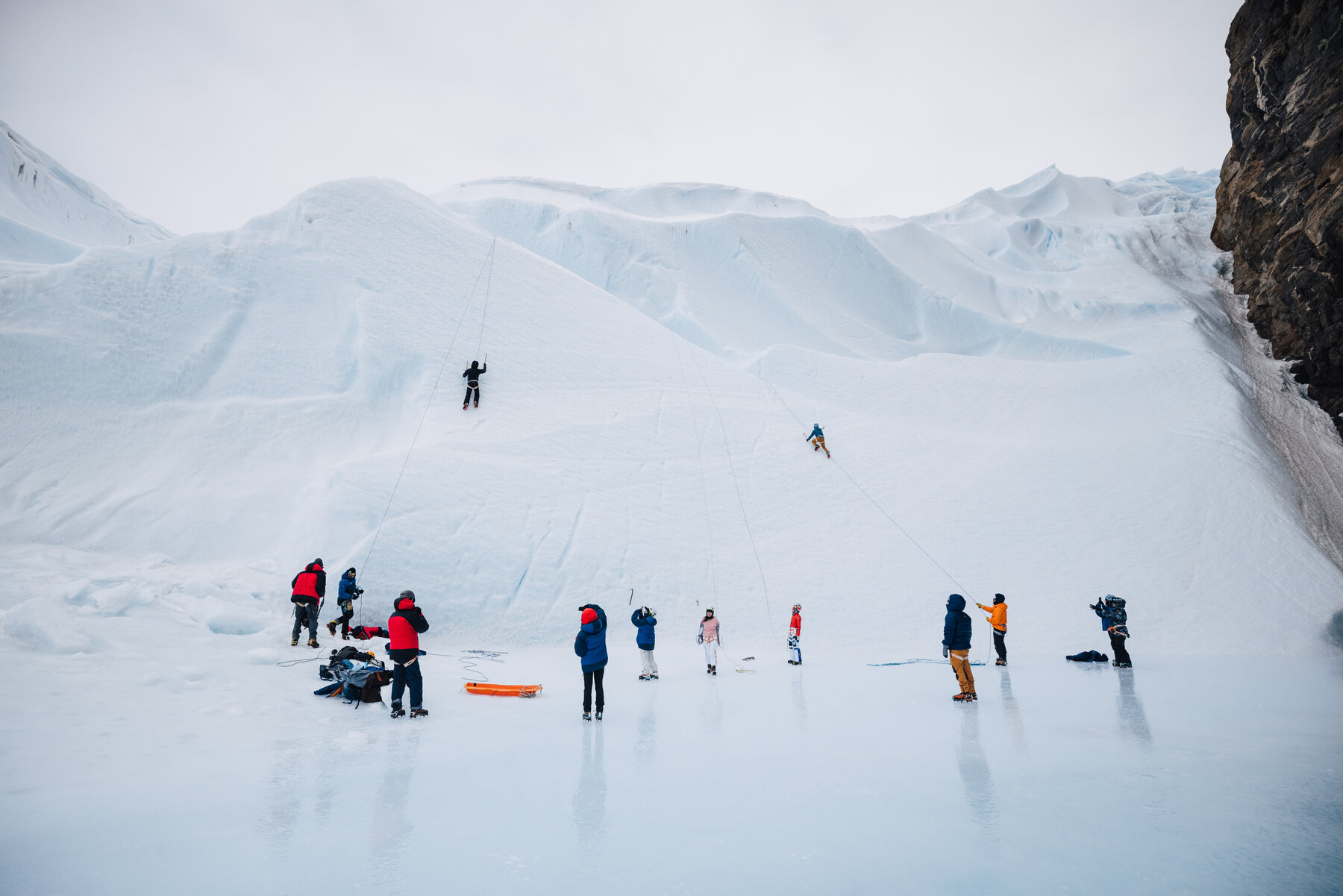 Wide scene of ice climbers on the blue and white ice face of the nunatak with belayers and gear spread across the plateau below