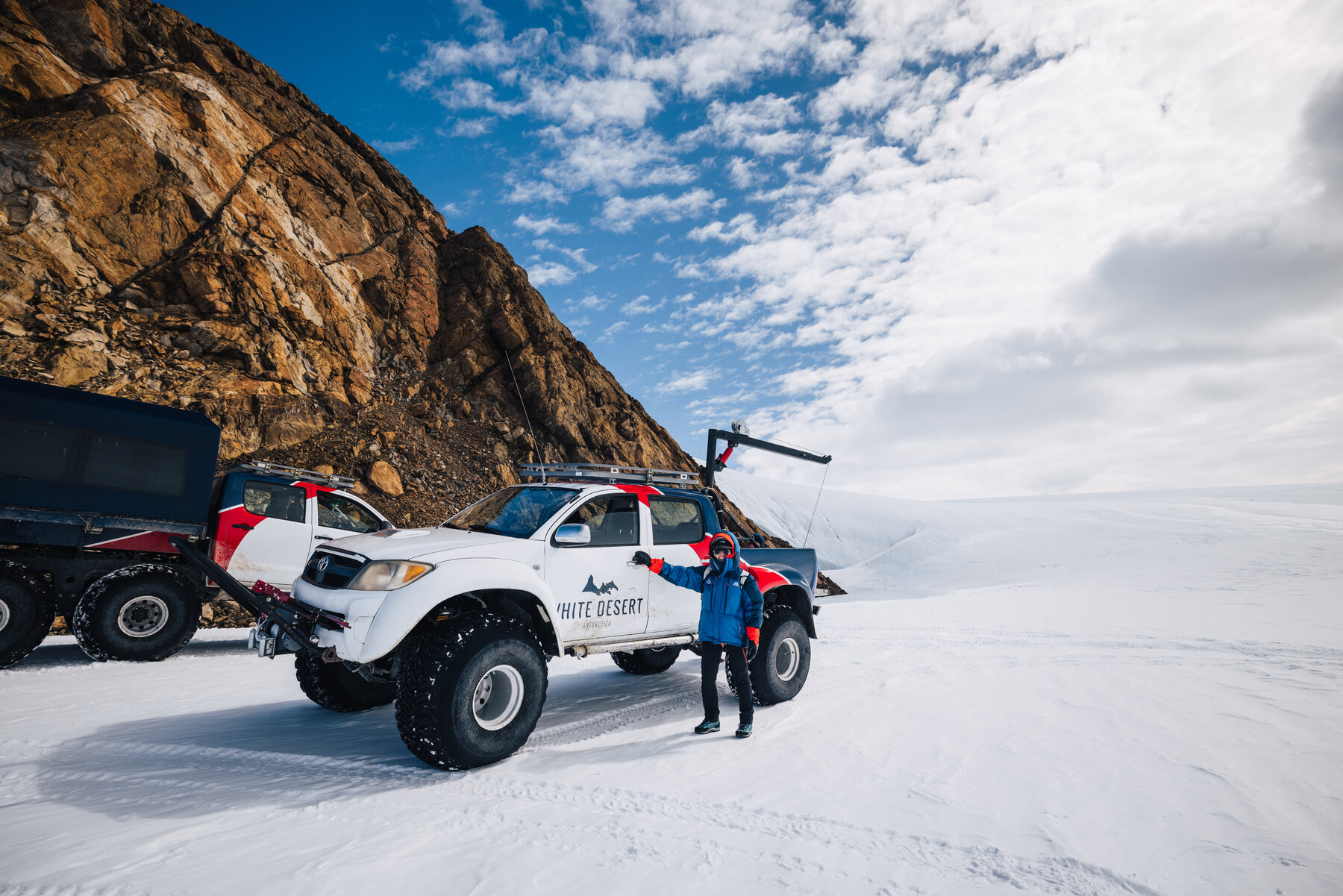 Jason in a blue parka beside a White Desert Arctic Trucks pickup on oversized tires, with a second expedition truck and rocky nunatak cliffs behind