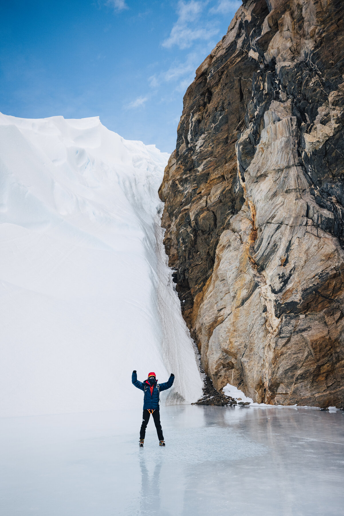 Climber in blue parka standing with arms raised at the base of a massive dark rock wall on the nunatak with snowdrifts piled against it
