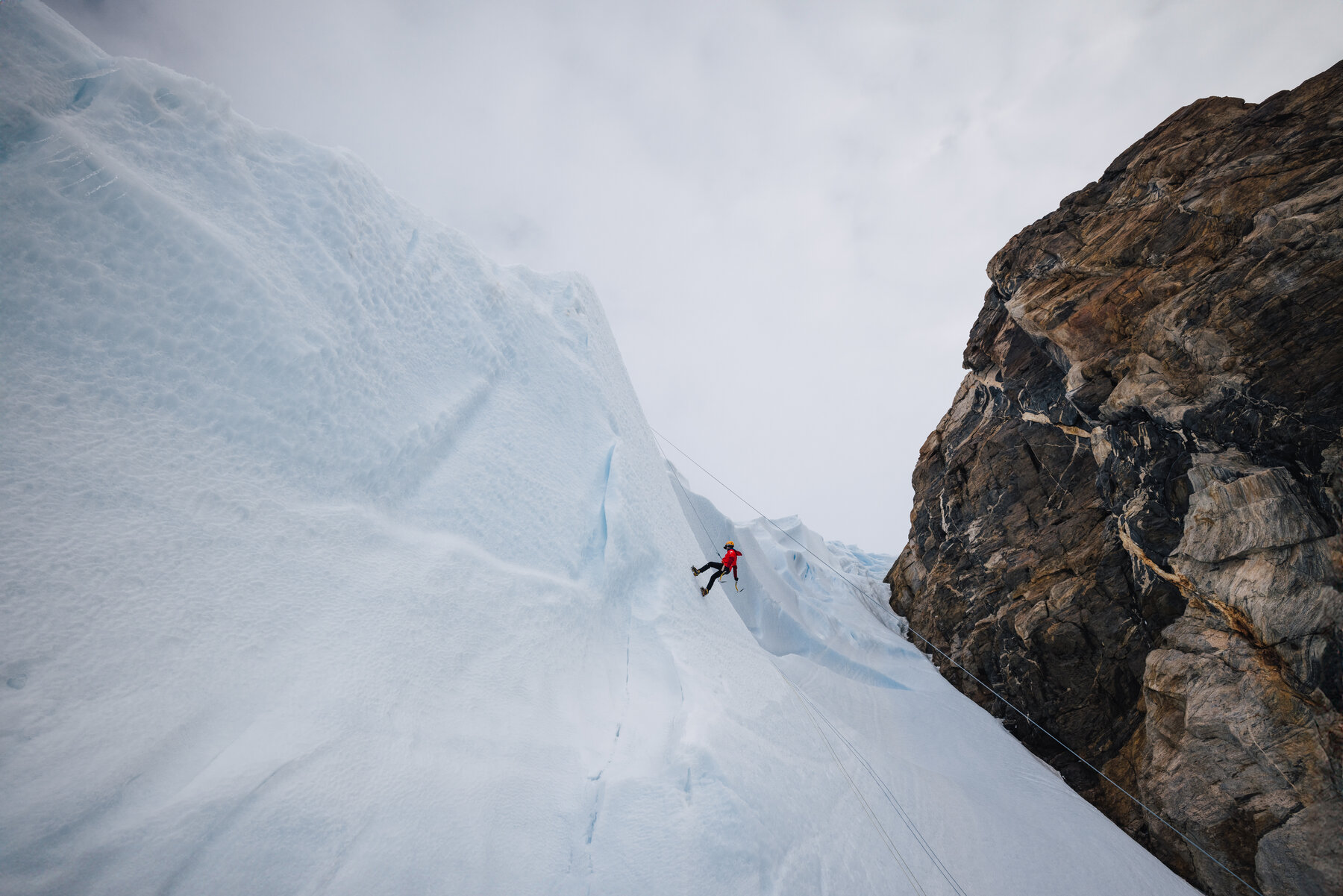 Lone climber in a red jacket rappelling down a massive blue and white glacial ice face beside a dark rock wall on the nunatak