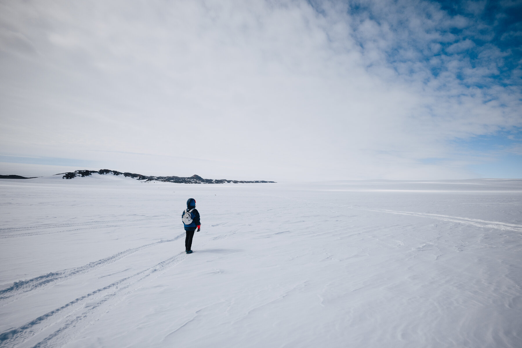 Lone figure walking across a vast empty Antarctic plateau toward distant rocky ridges under a high scattered-cloud sky