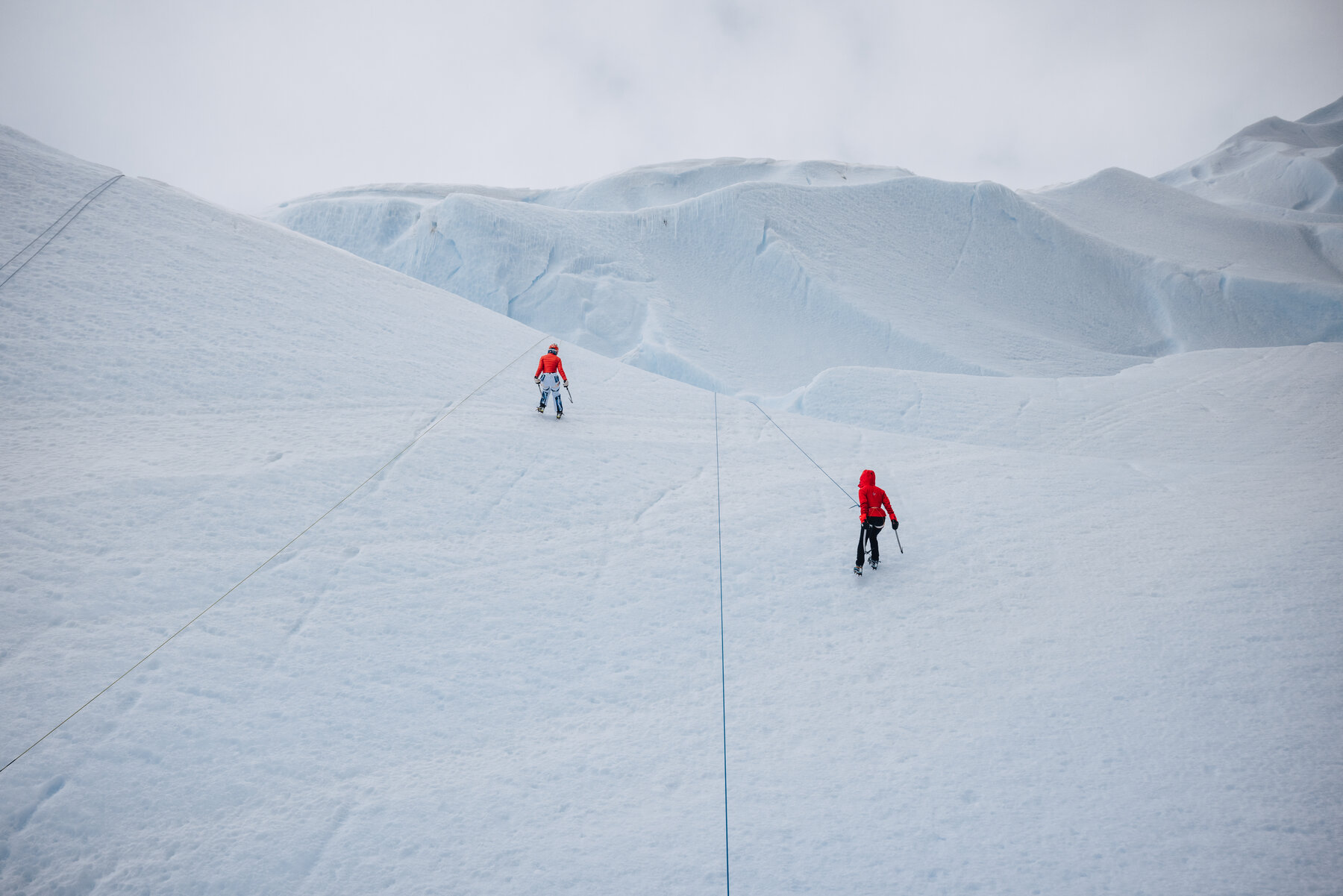 Two climbers in red jackets walking a snow ridge with trailing rope and distant ice cliffs on the horizon
