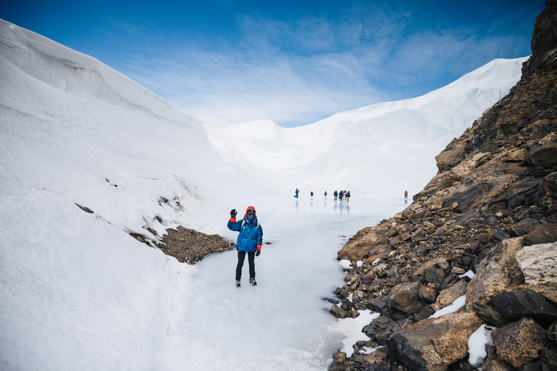 Climber in blue parka walking through a snow canyon flanked by drifts and rocky outcrops with group members ahead in the distance