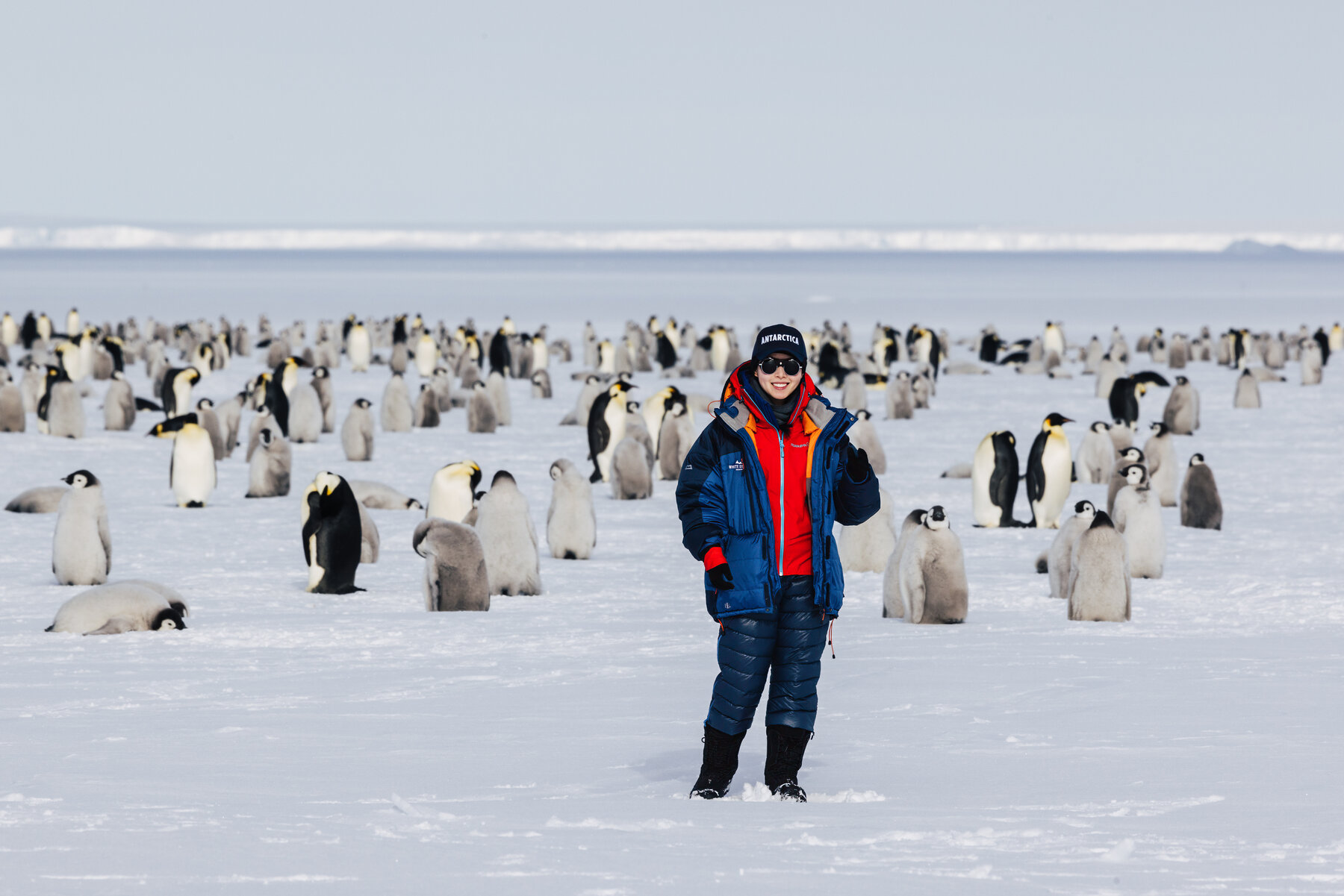 Grace in a blue parka and red jacket standing in the middle of the Emperor penguin colony with a flat ice horizon behind