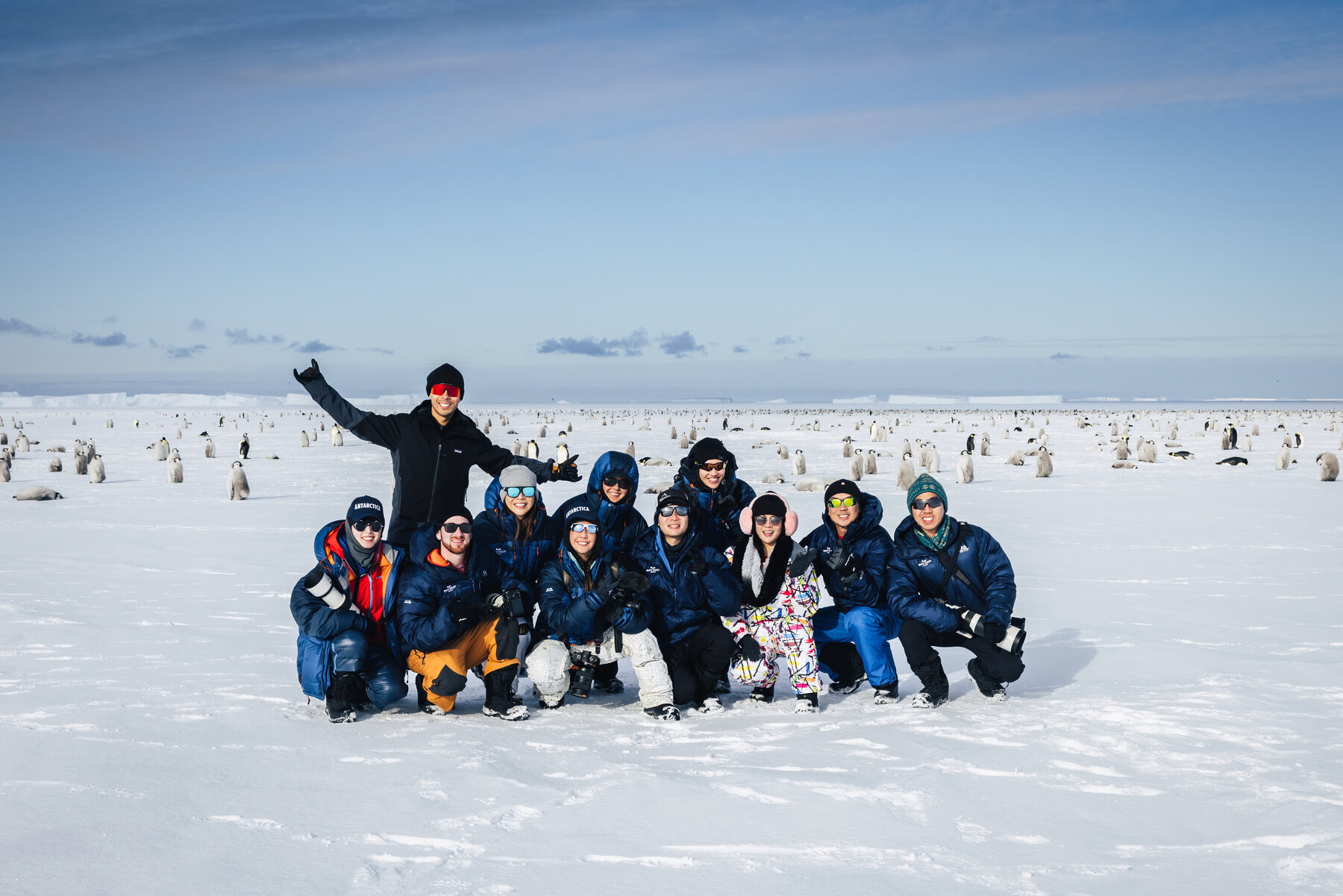 Group photo of ten travelers posed together on the sea ice with the Emperor penguin colony spread behind them