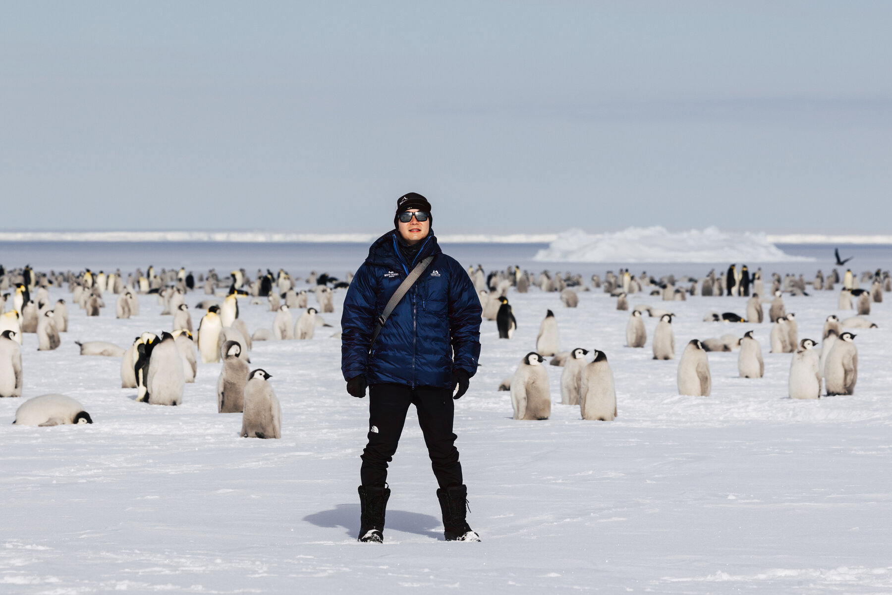 Jason in a blue parka standing confidently among the Emperor penguin colony with adults and chicks spread on both sides