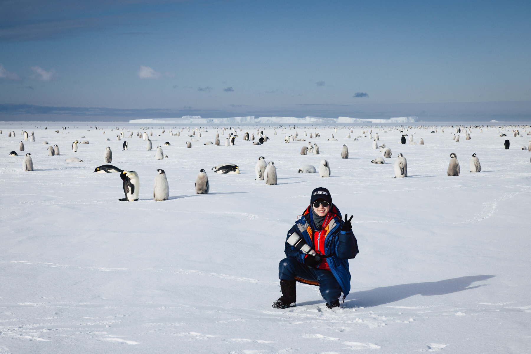 Jason crouching in a dark parka making a peace sign with the Emperor penguin colony spread across the ice behind him