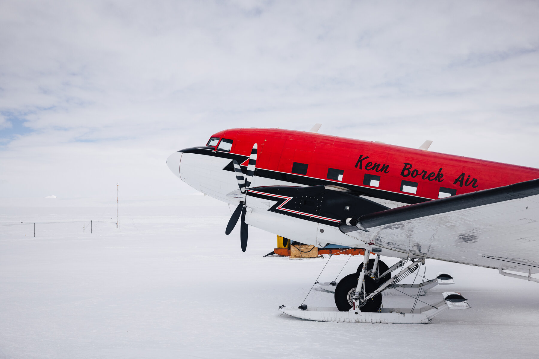 Red and white Kenn Borek Air DC-3 aircraft with ski landing gear parked on the ice at Atka Bay