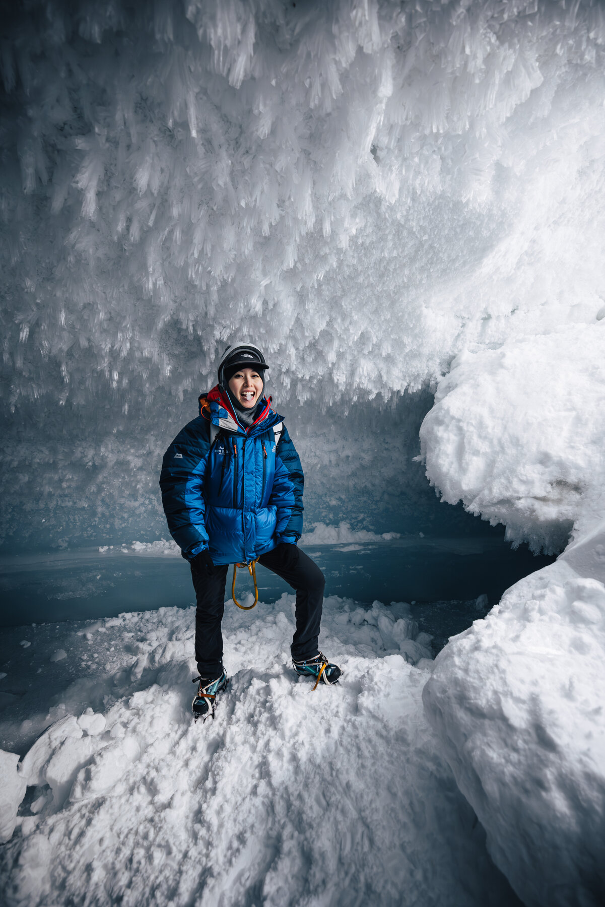 Grace in a blue parka and crampons standing inside the ice cave smiling at the camera with crystal walls behind her