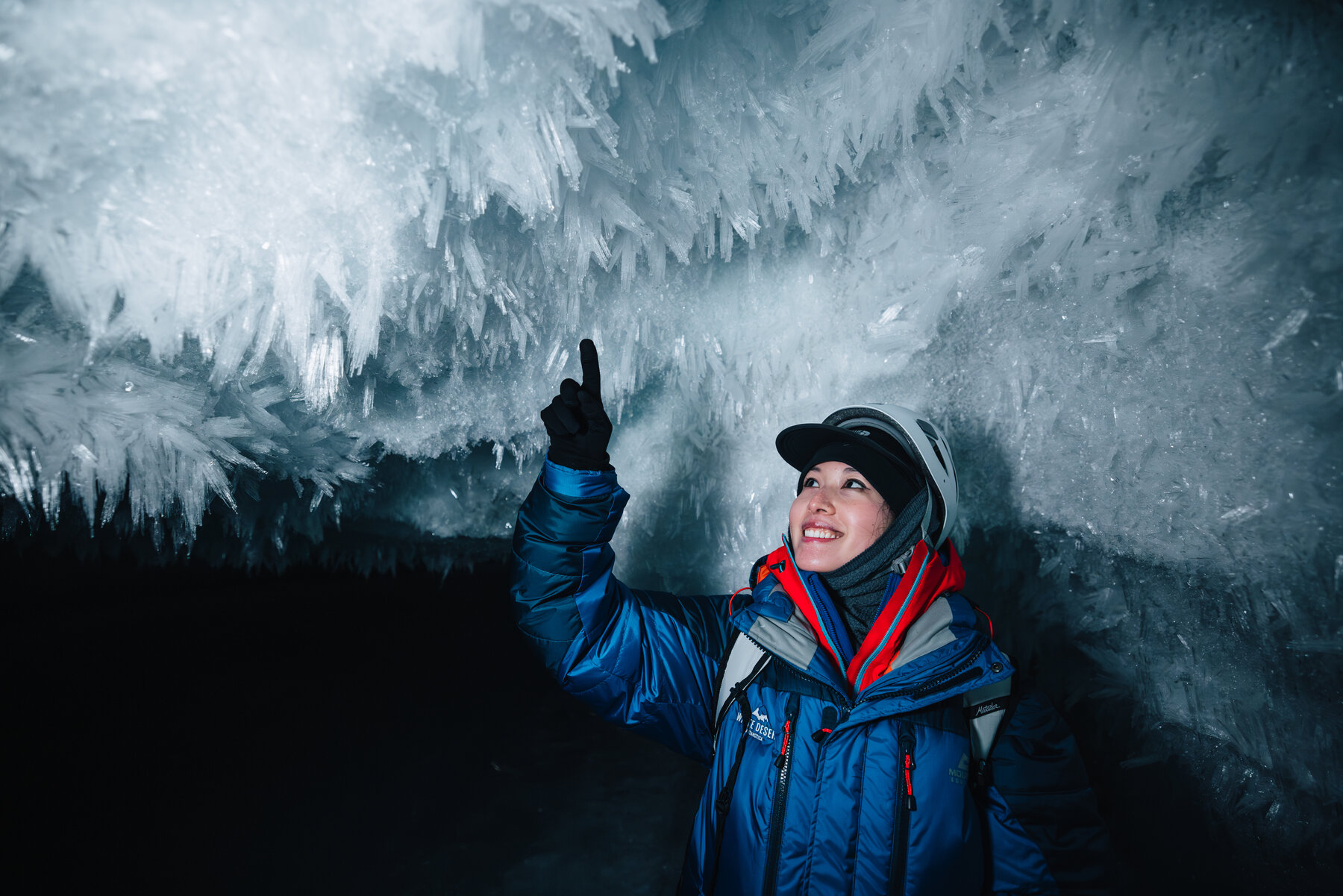 Grace in a blue parka smiling and pointing up at the crystal ice formations on the cave ceiling