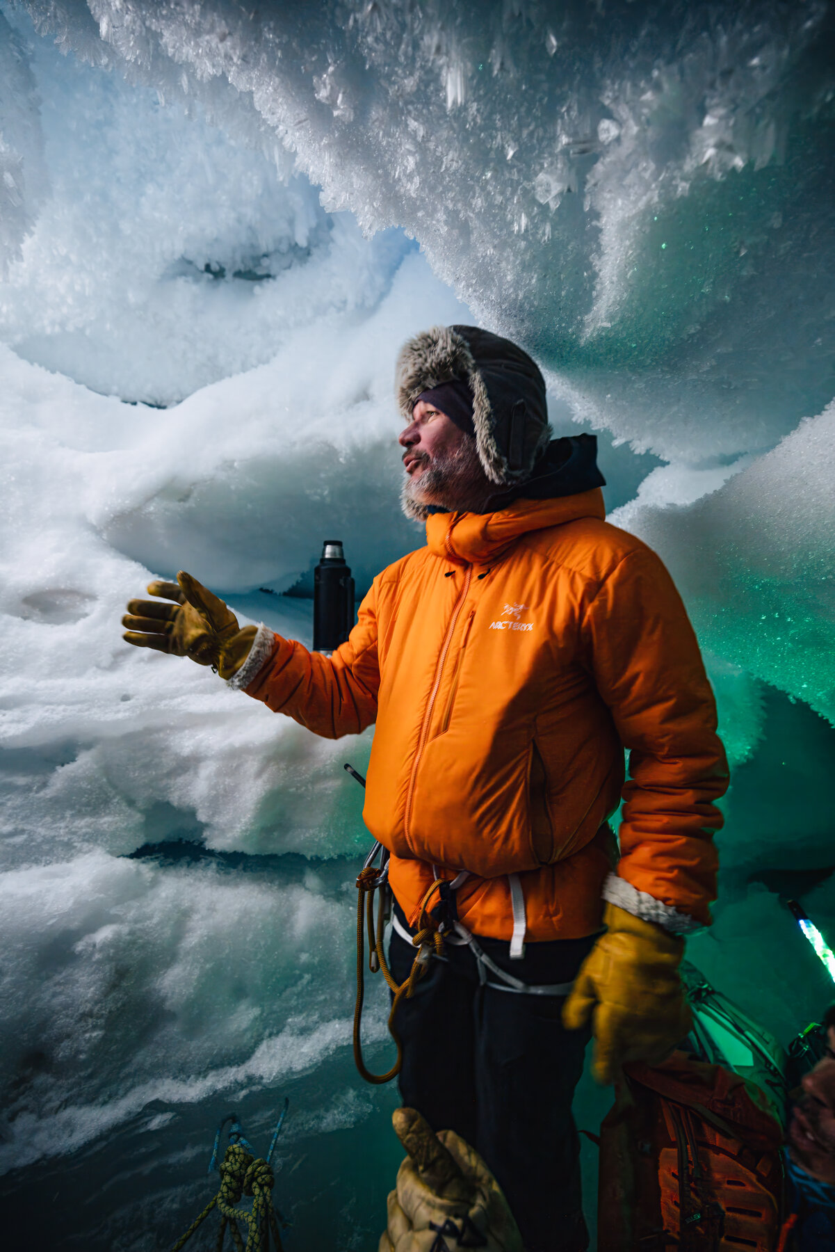 Guide in an orange parka and fur-lined hat gesturing mid-explanation inside the ice cave with green-tinted ice walls behind him