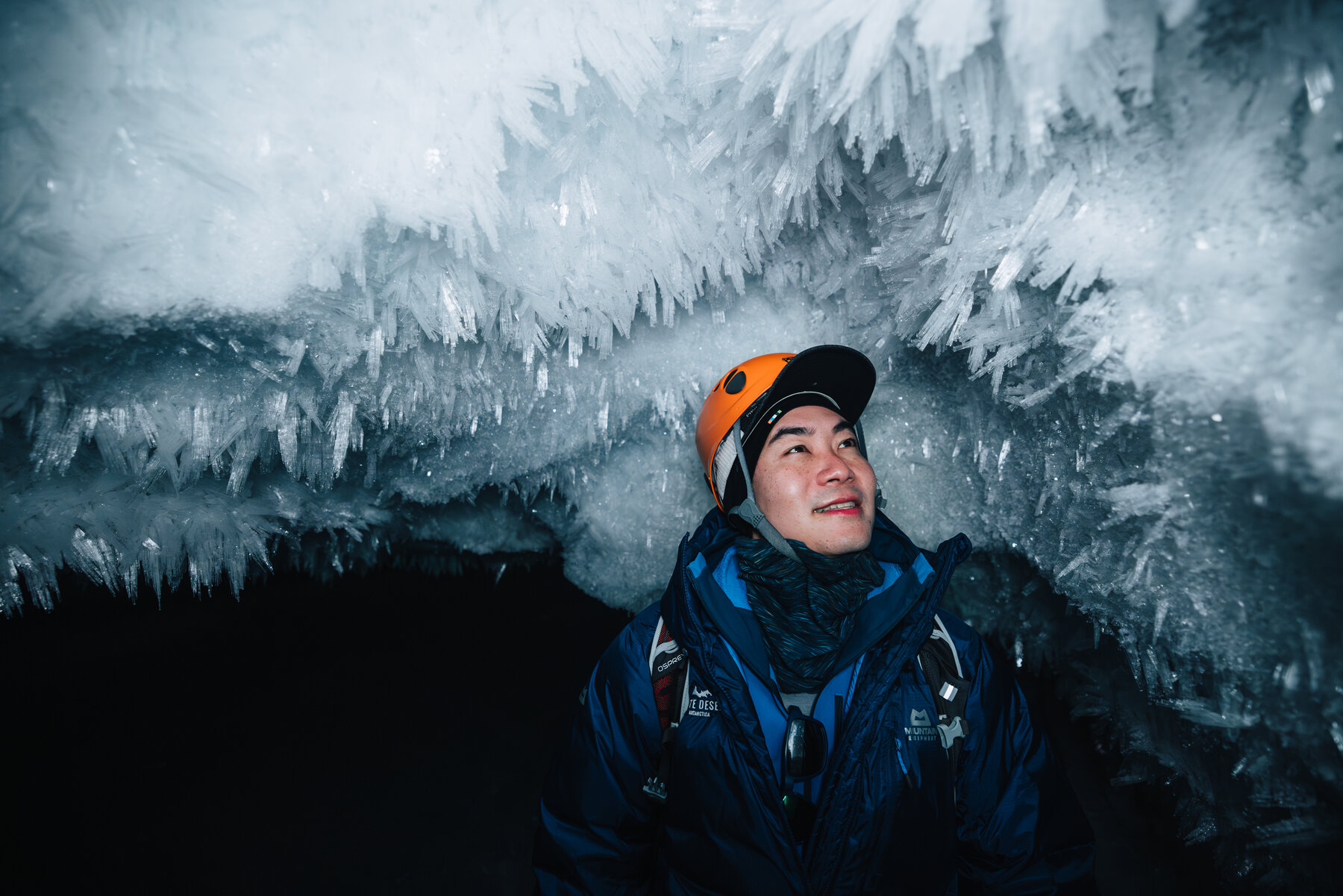 Jason in an orange climbing helmet and blue parka looking up at the crystal-encrusted ceiling inside the ice cave