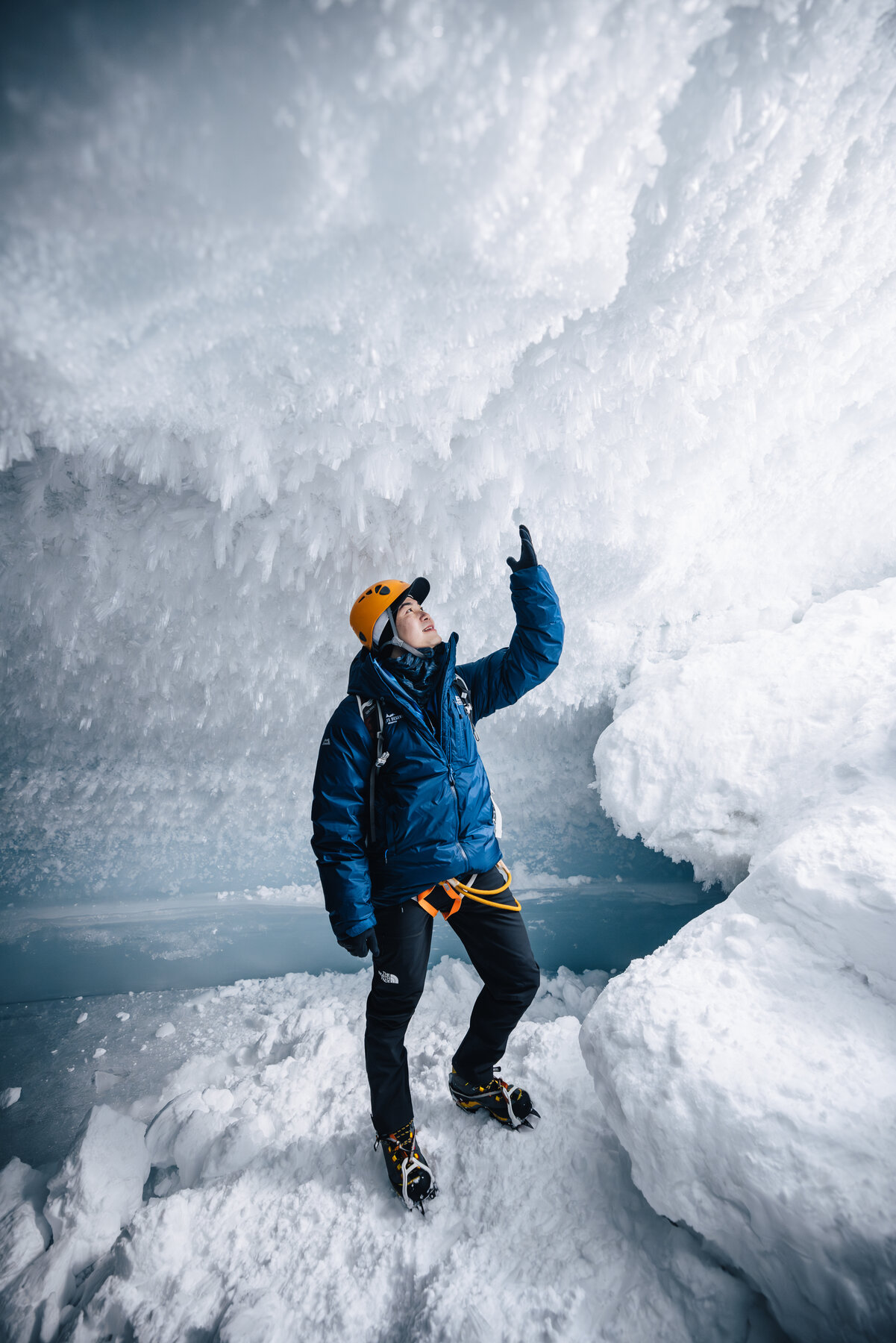 Jason in a blue parka and orange helmet reaching up to touch the crystal ice ceiling of the cave with crampons on his boots