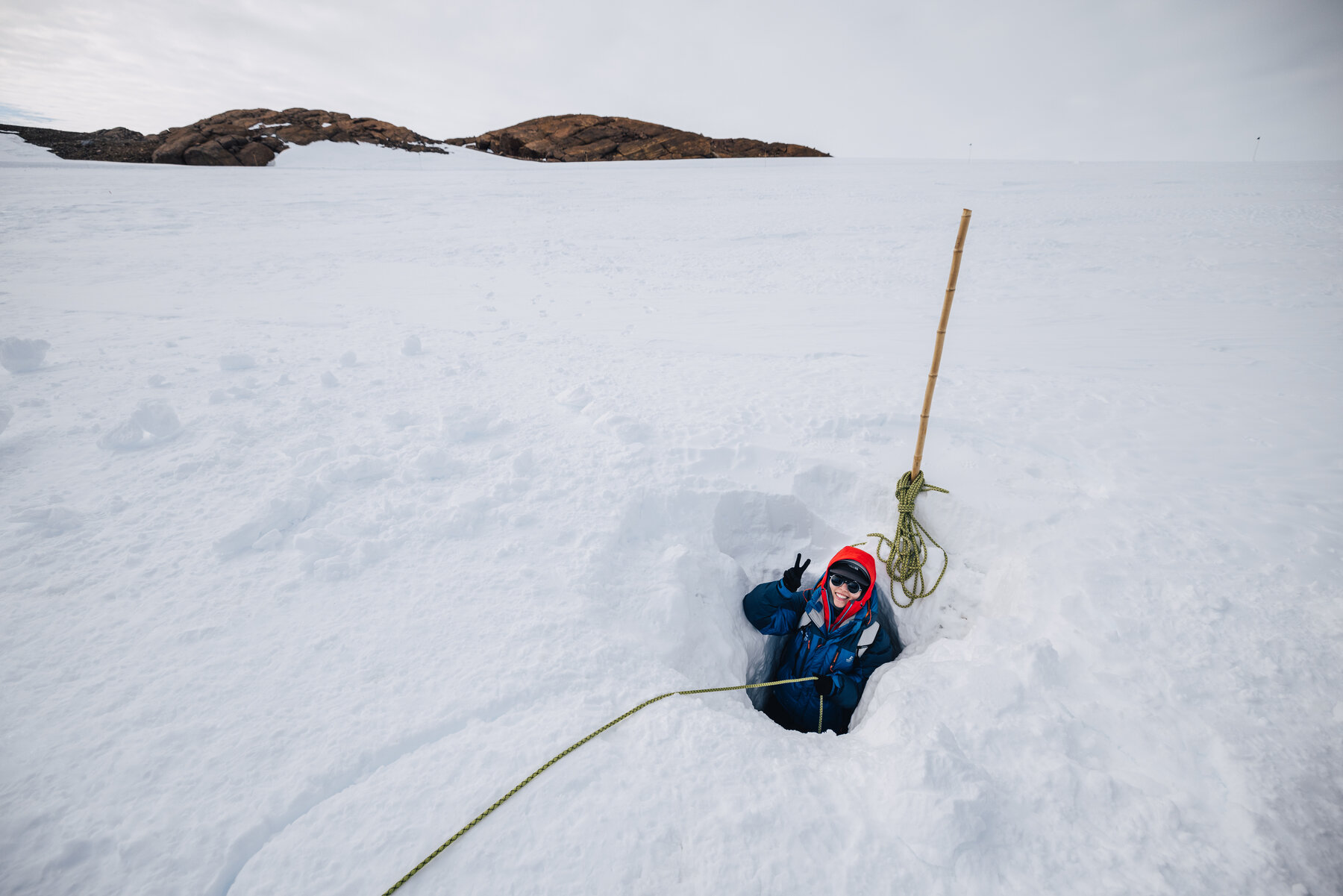 Climber in a red hat sitting waist-deep in a snow hole at the cave entrance with a climbing rope trailing across the ice toward distant nunataks