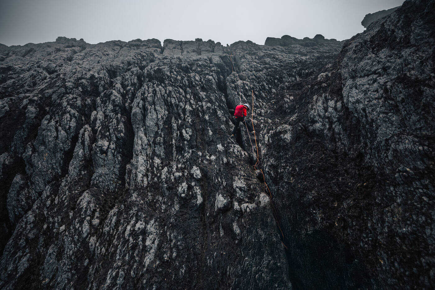 Climber in a red jacket on a fixed rope high on a dark, fluted limestone face, seen from above