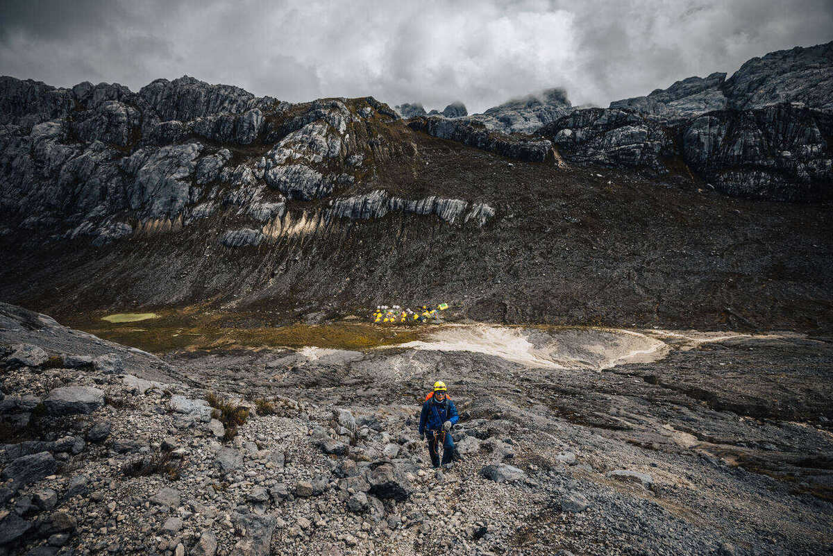 Climber in blue jacket and helmet on rock field with dramatic limestone walls behind